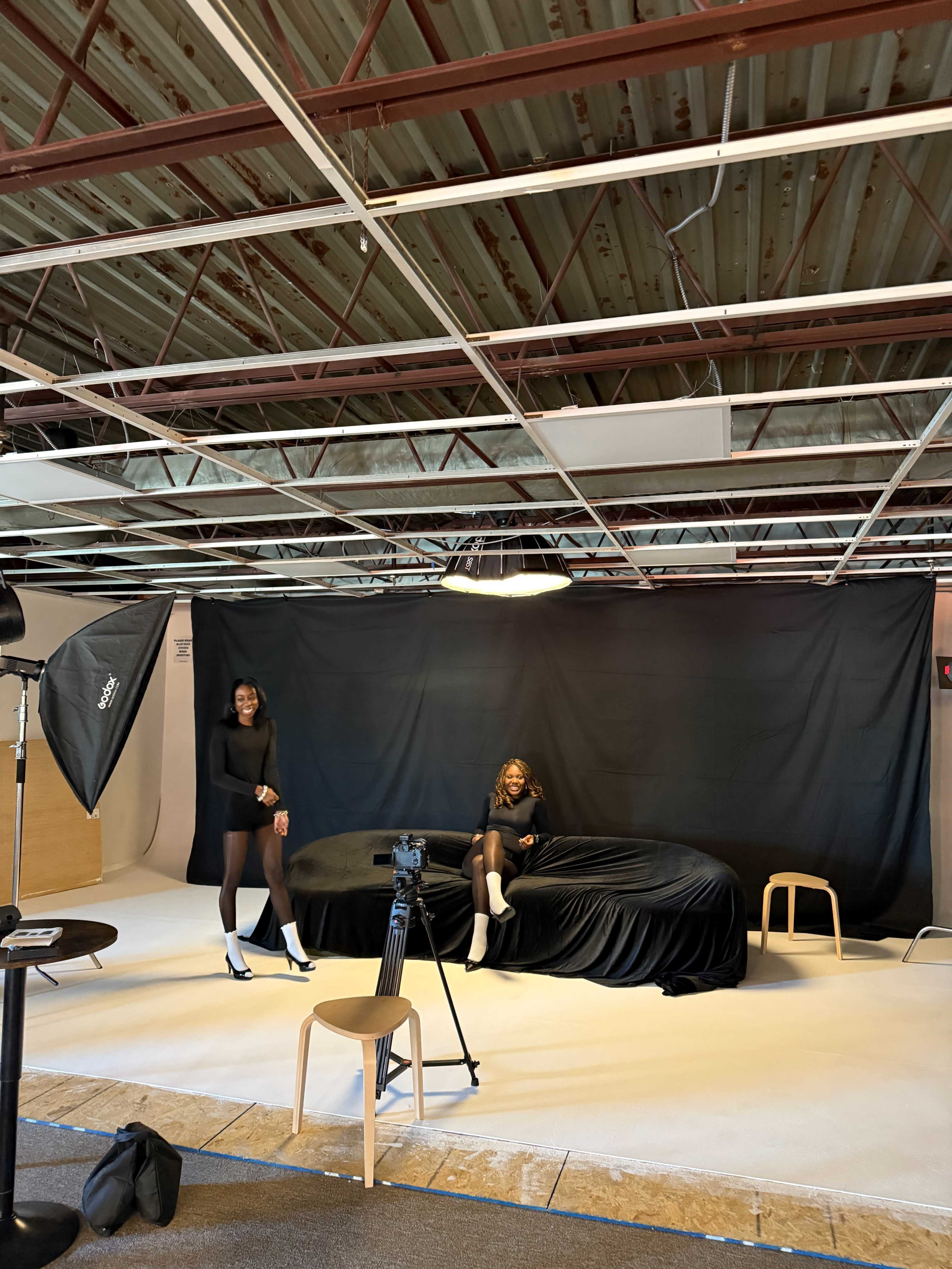 Two women pose in a studio setting with a black backdrop, while a camera and softbox lighting equipment are set up nearby.
