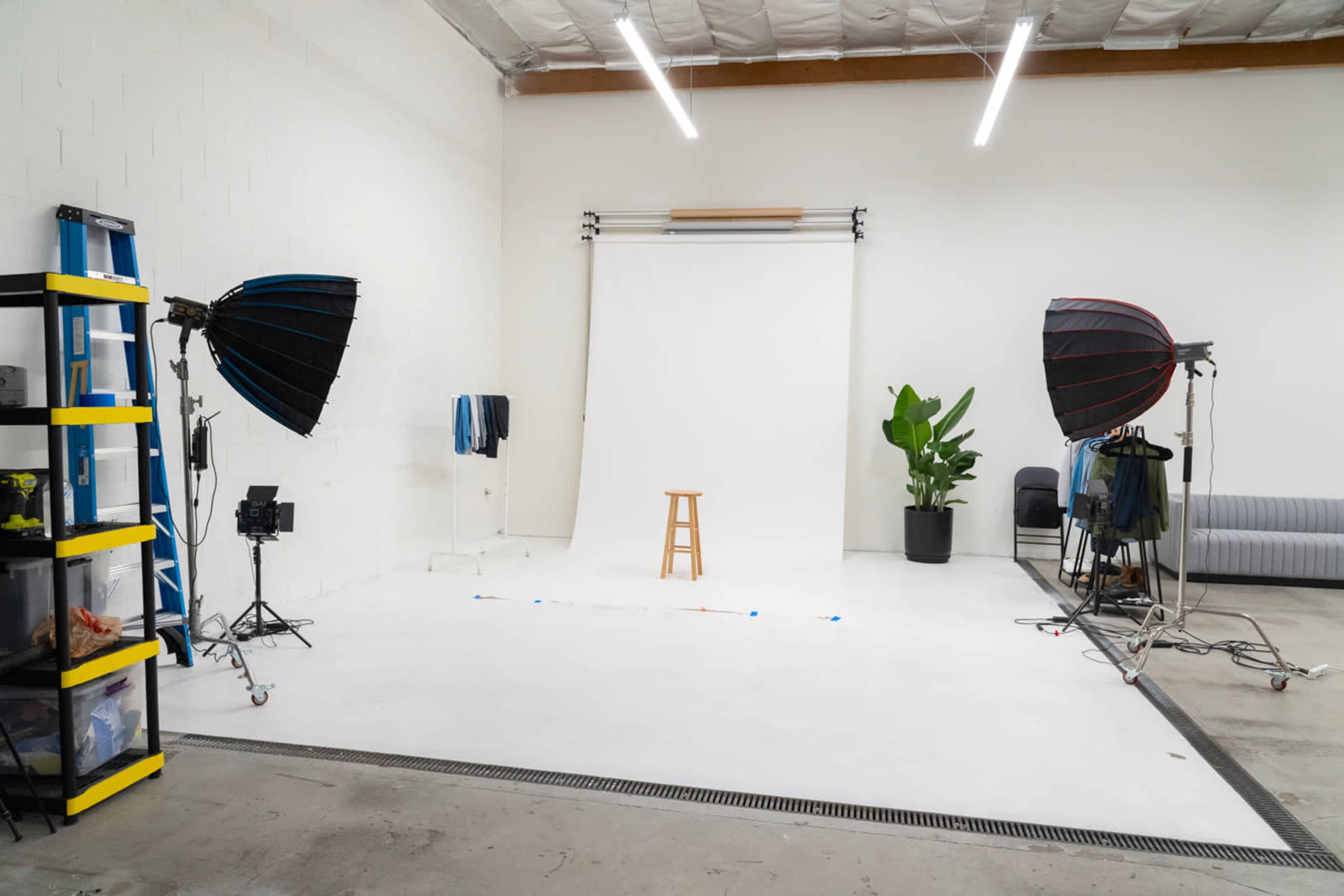 A photography studio with a white backdrop, a wooden stool in the center, two softbox lights on either side, and various equipment arranged around the space.