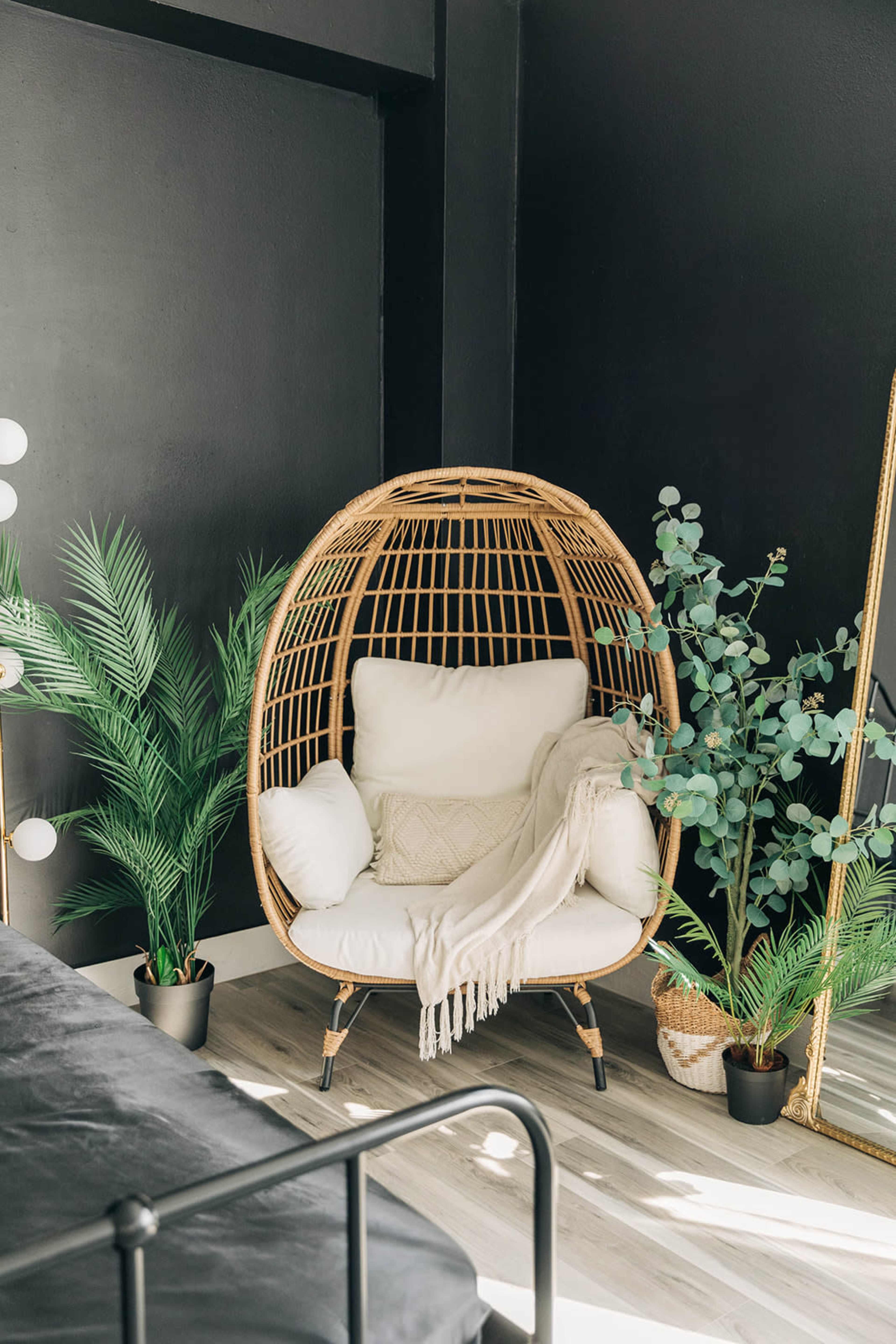 A rattan chair with white cushions is positioned in a corner surrounded by potted plants, against a black wall and near a gold-framed mirror.