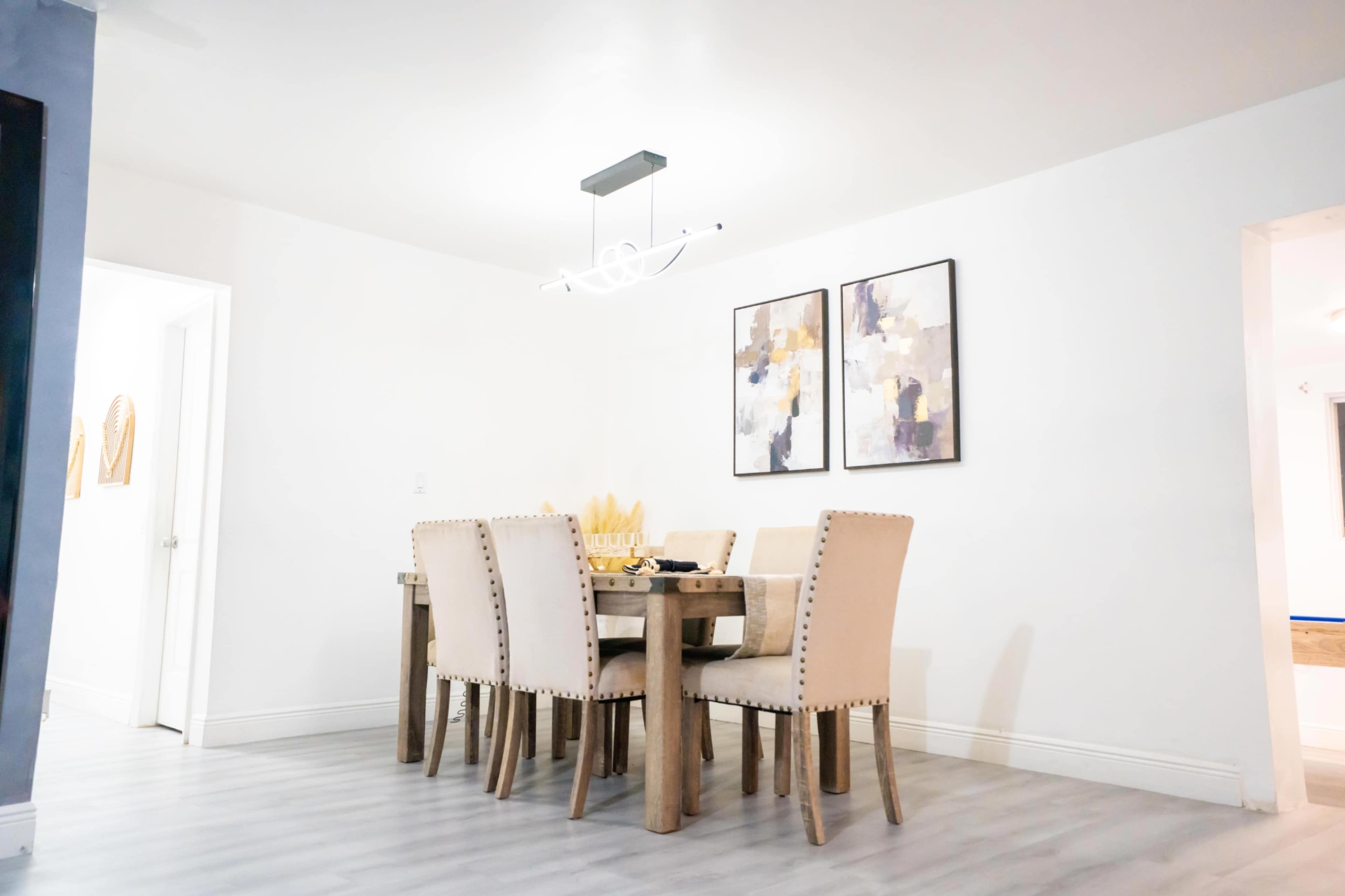 A dining area features a wooden table surrounded by six upholstered chairs, with two framed artworks on the wall above.