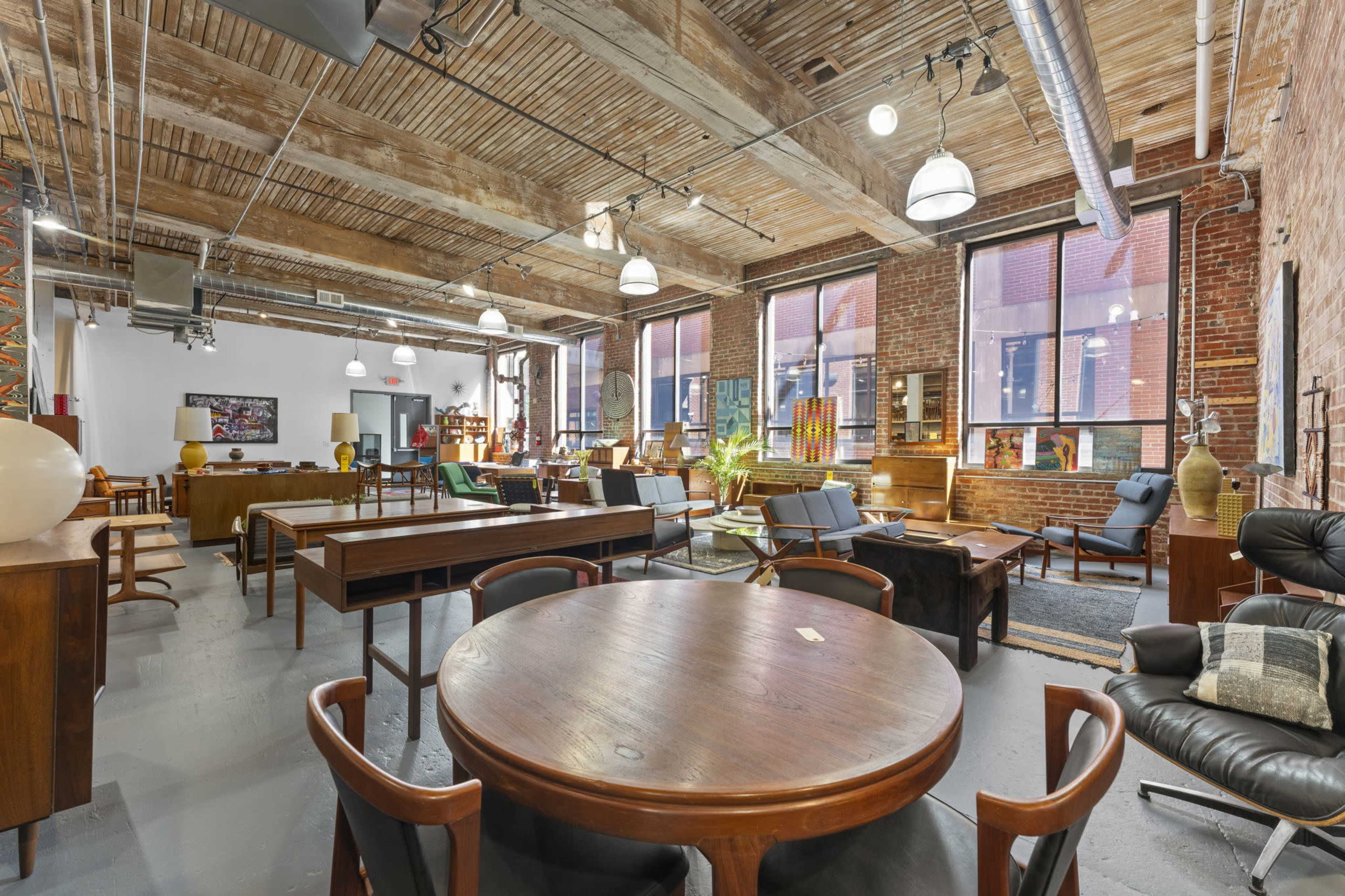 A spacious interior filled with various mid-century furniture pieces, featuring large windows, exposed brick walls, and wooden beams.