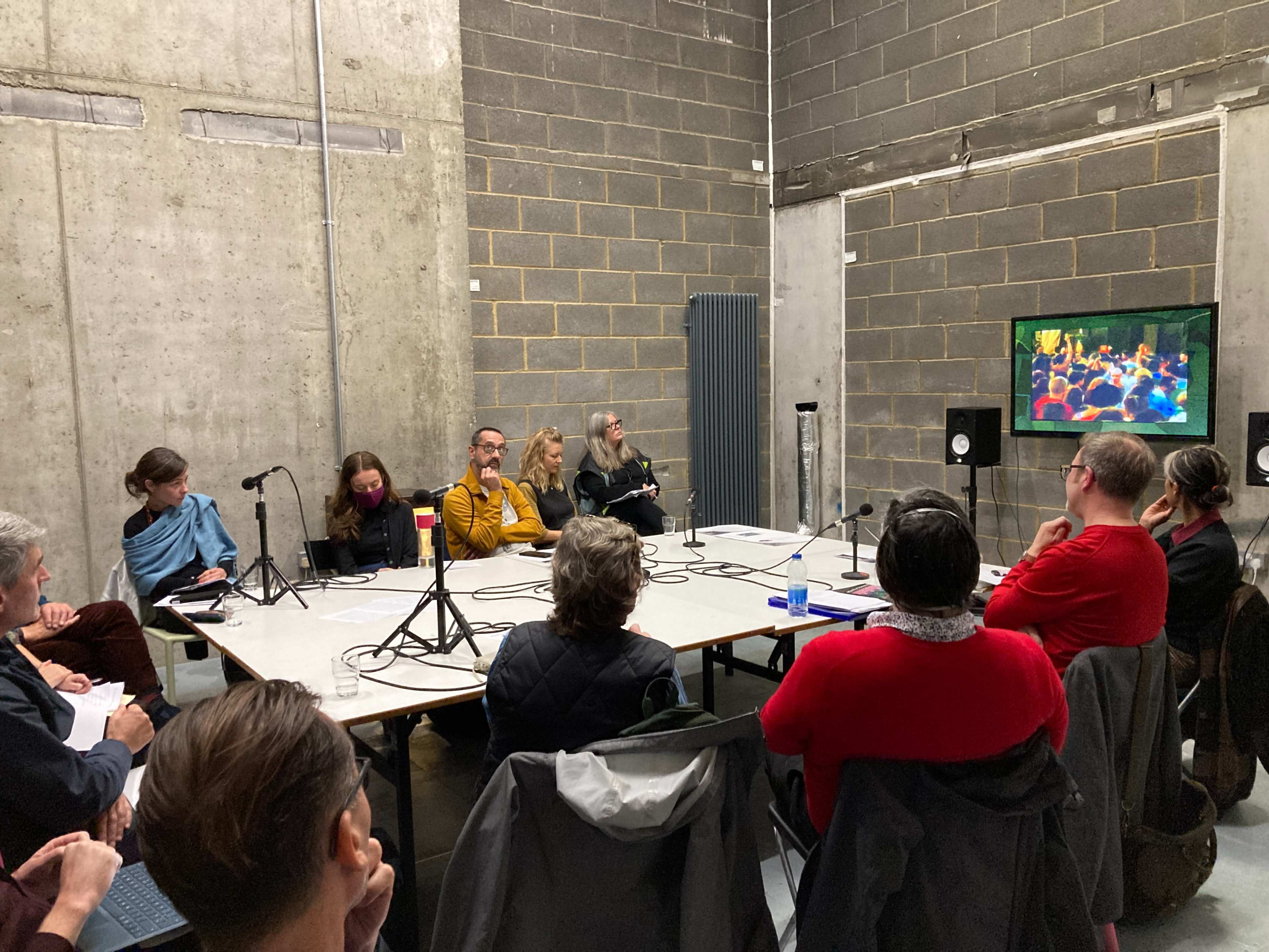 A group of people sits around a table in a gray-walled room, watching a video presentation on a screen.