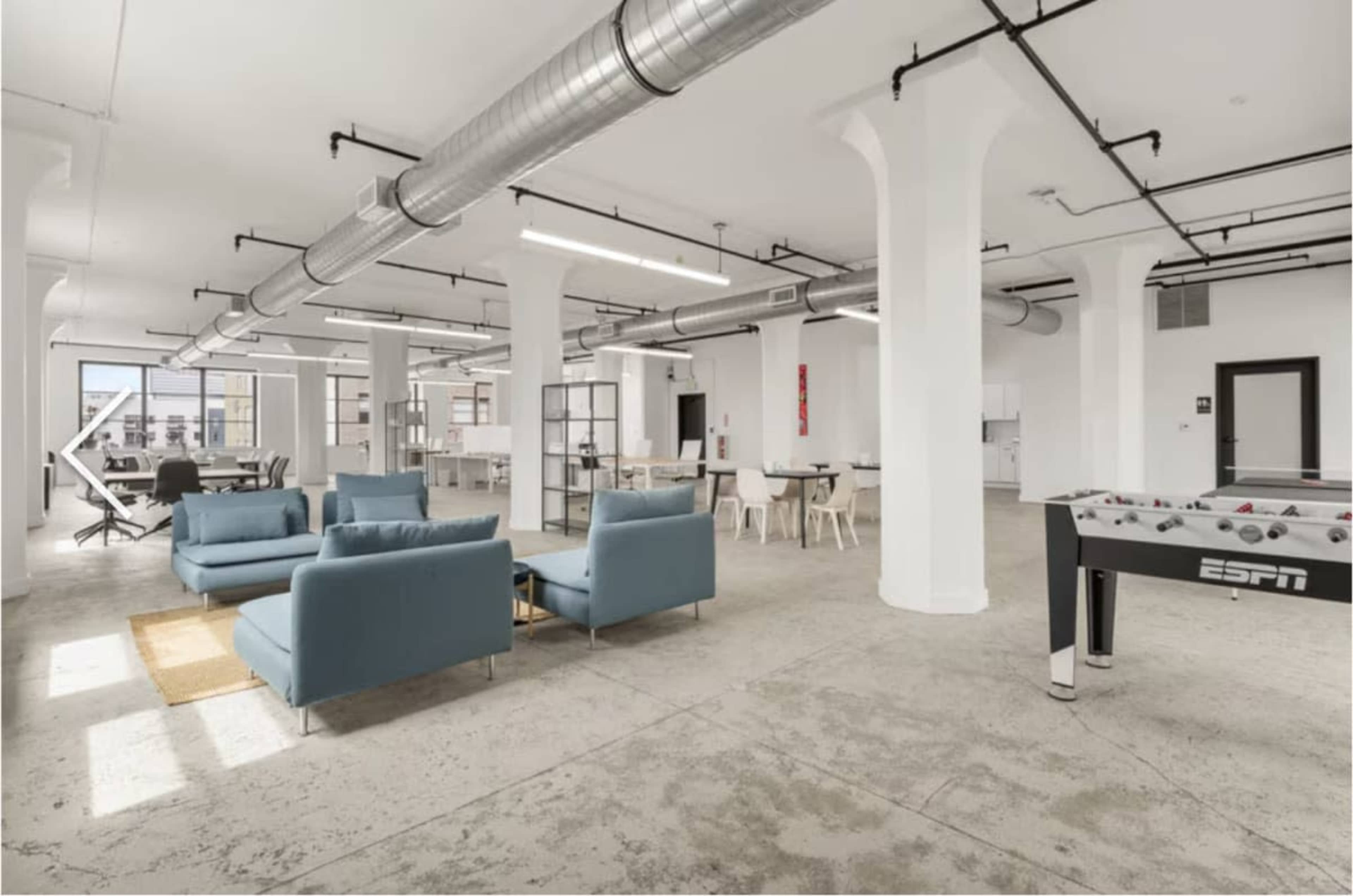 The image shows a spacious, modern office area featuring light-colored furniture, large windows, and a foosball table.