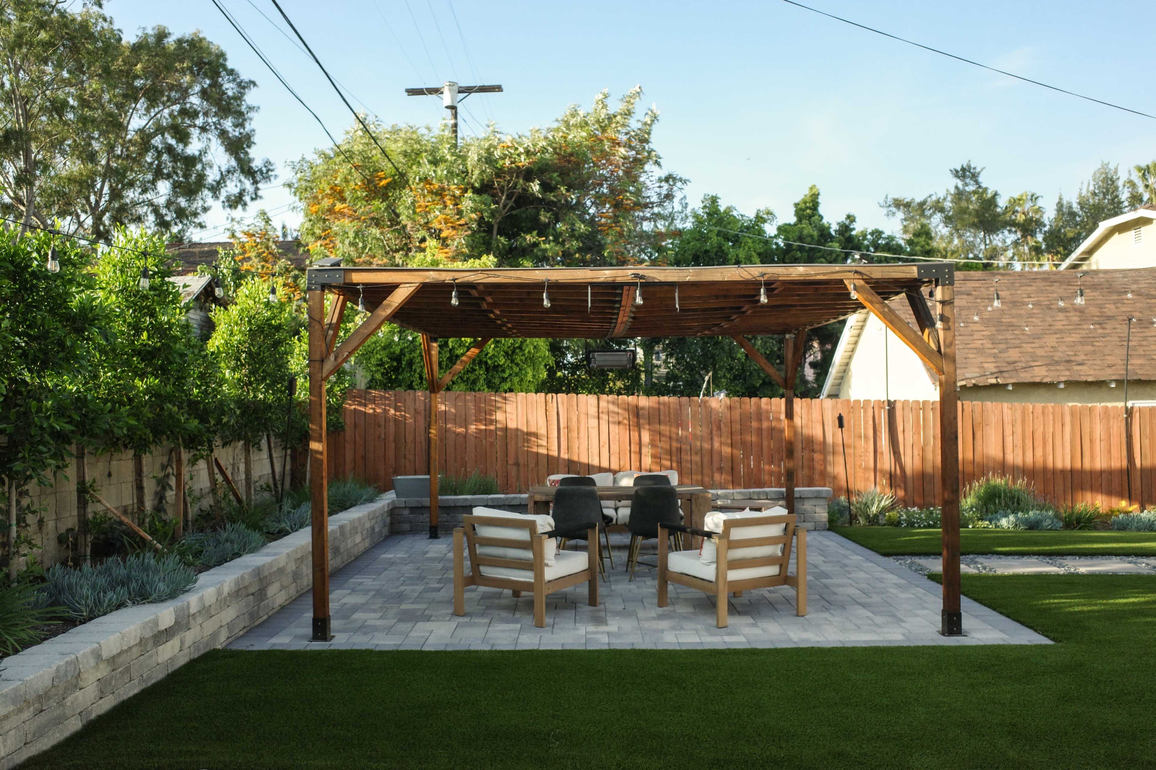 The image shows a backyard with a wooden pergola over a stone patio, furnished with four chairs and a table, surrounded by greenery and a wooden fence.