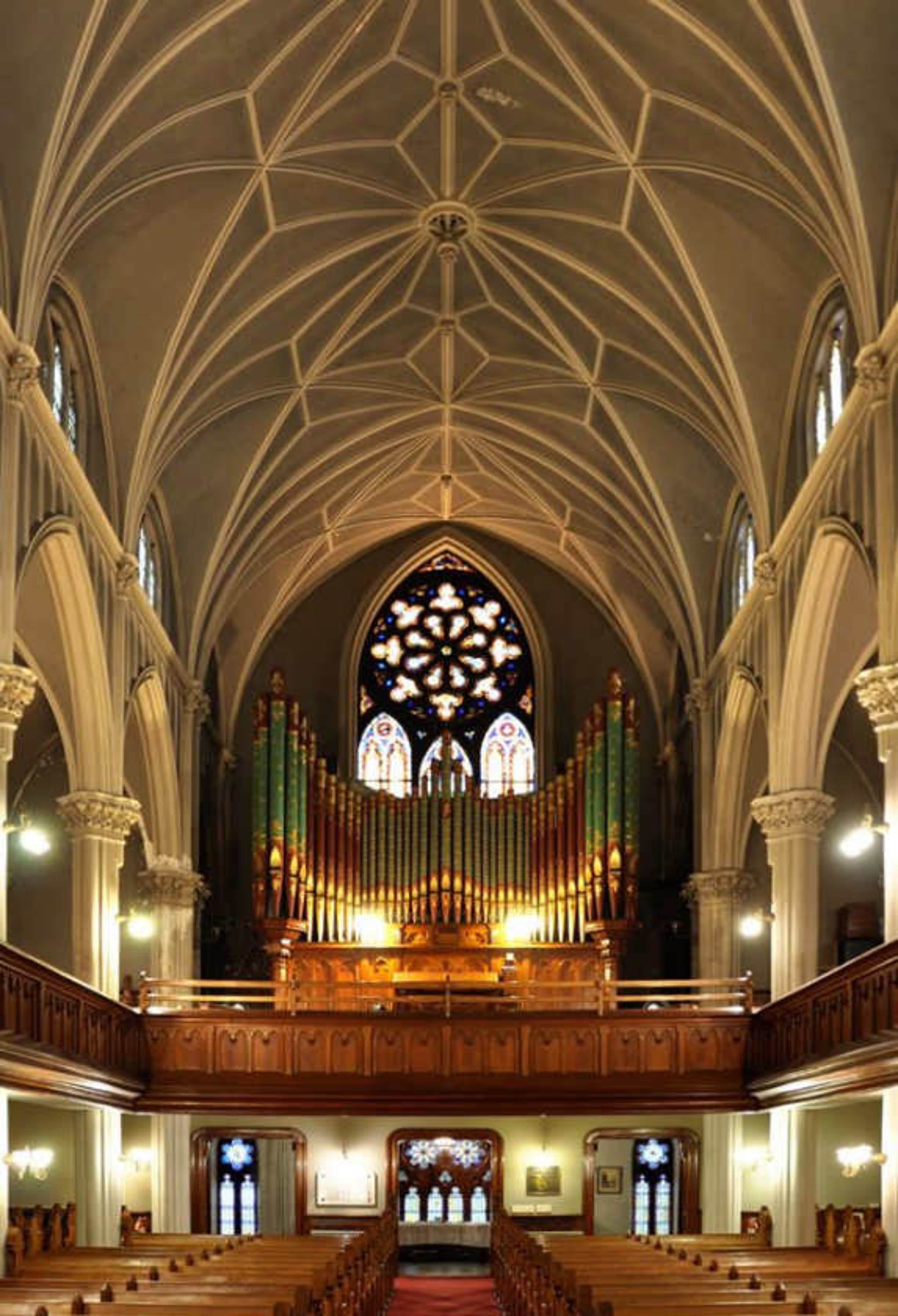 The image shows the interior of a church featuring a high vaulted ceiling, ornate columns, and a large stained glass window above an organ.