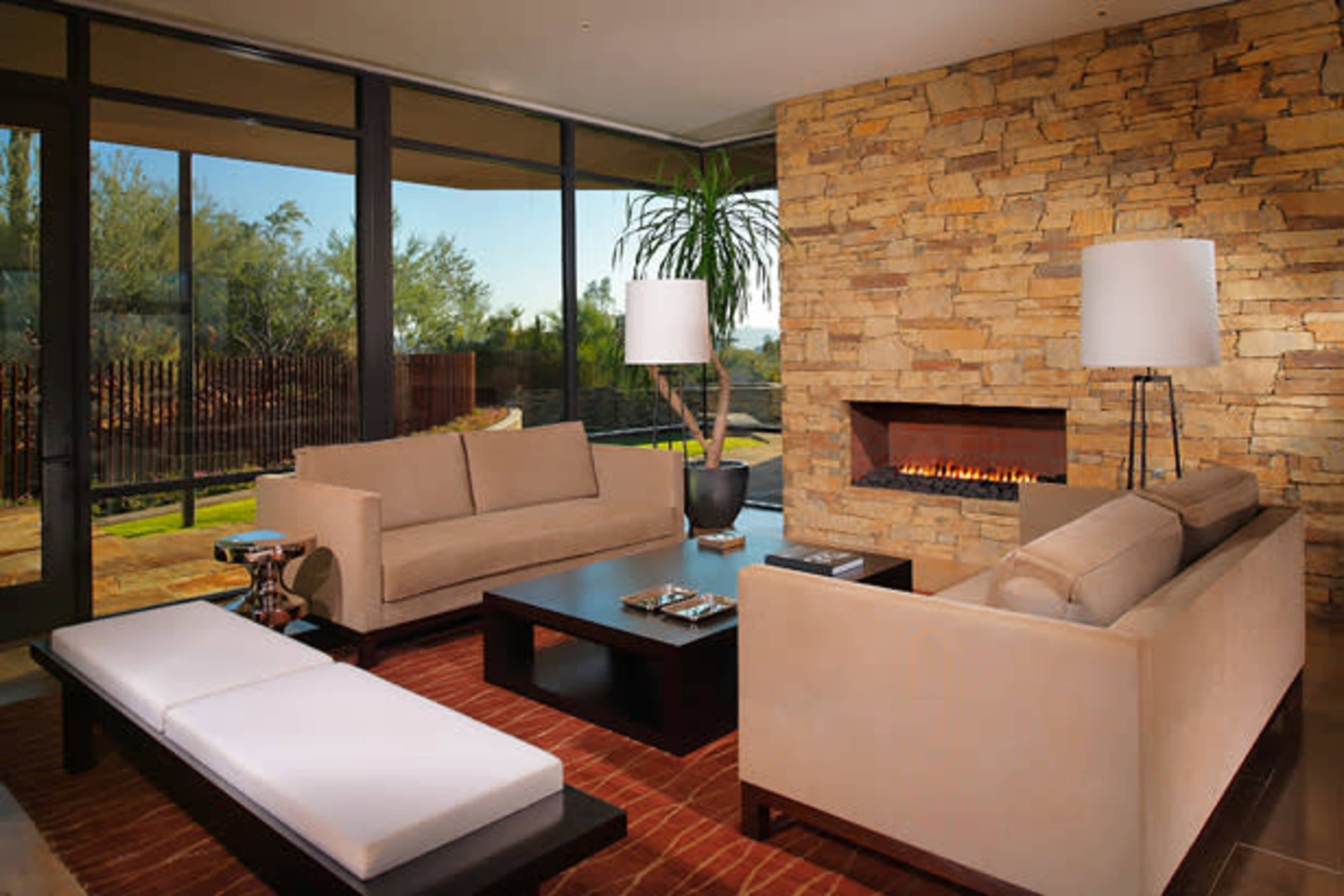 A modern living room features a stone fireplace, large windows, and contemporary furniture arranged around a coffee table.