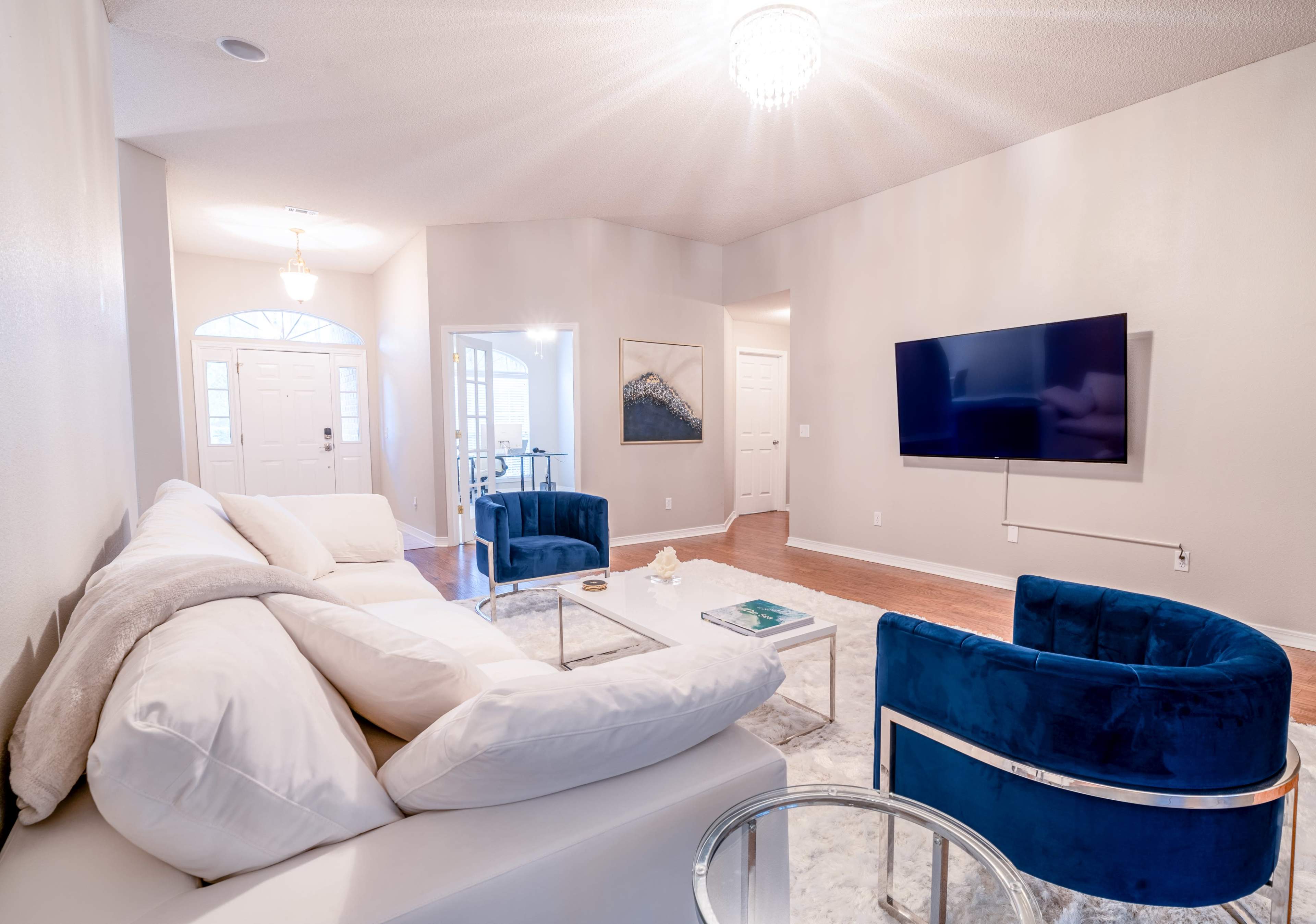 A well-lit living room featuring a white sectional sofa, two blue armchairs, a glass coffee table, and a wall-mounted television.