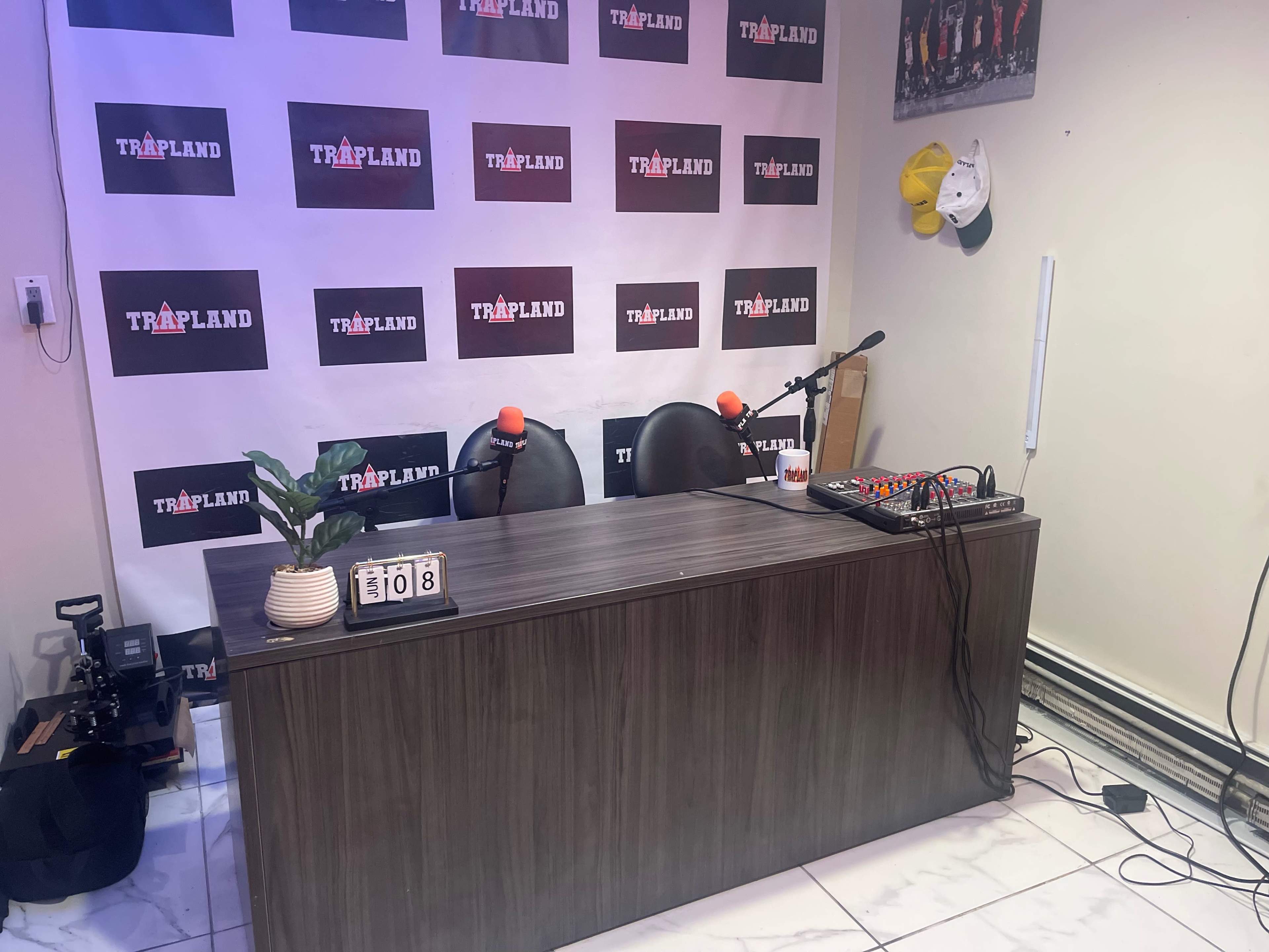 A small media studio setup with a wooden desk, two chairs, microphones, and a control panel, against a background featuring the logo "TRAPLAND."