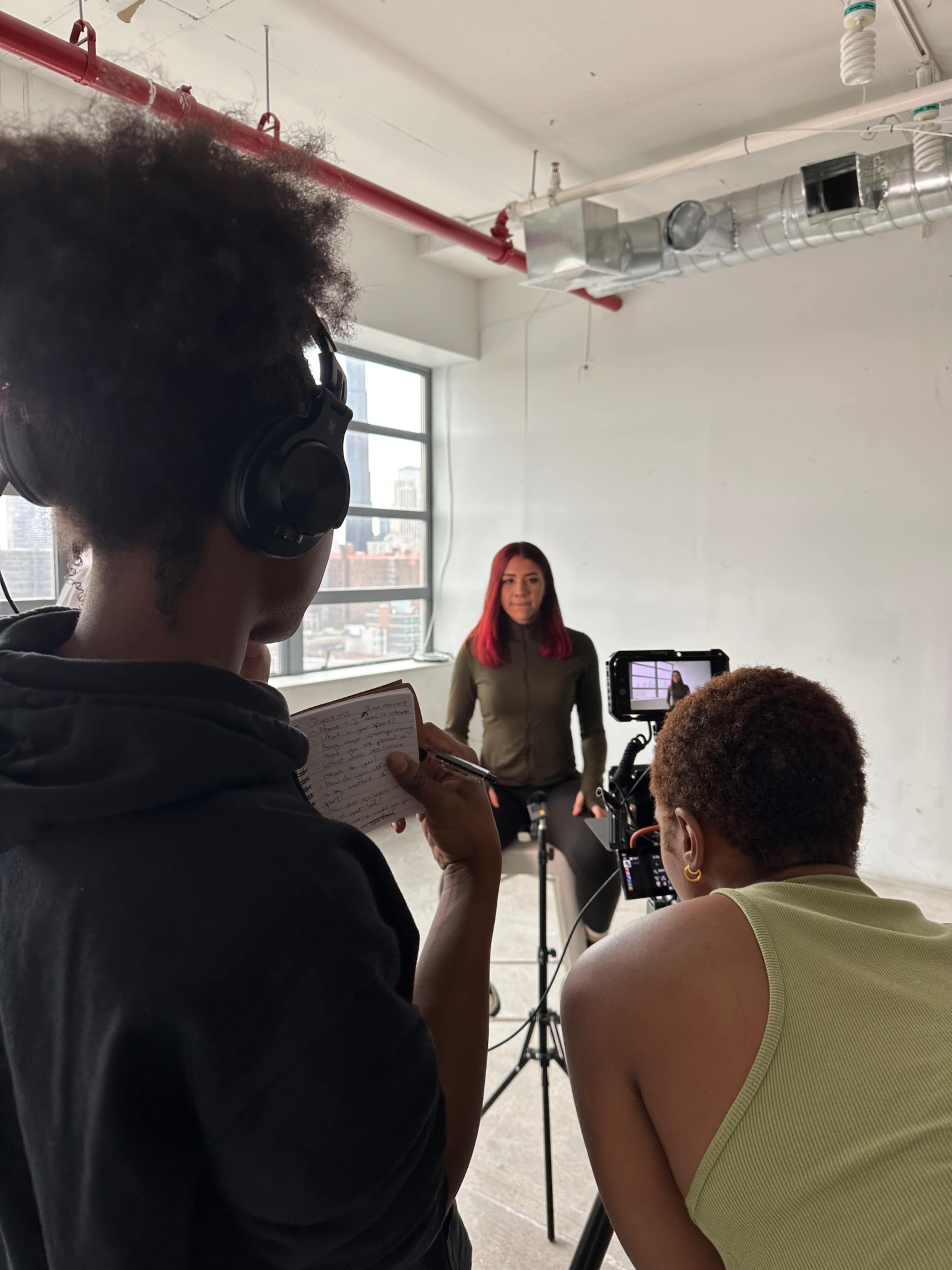 A woman with red hair sits on a stool while two crew members prepare to film her in a well-lit industrial space.
