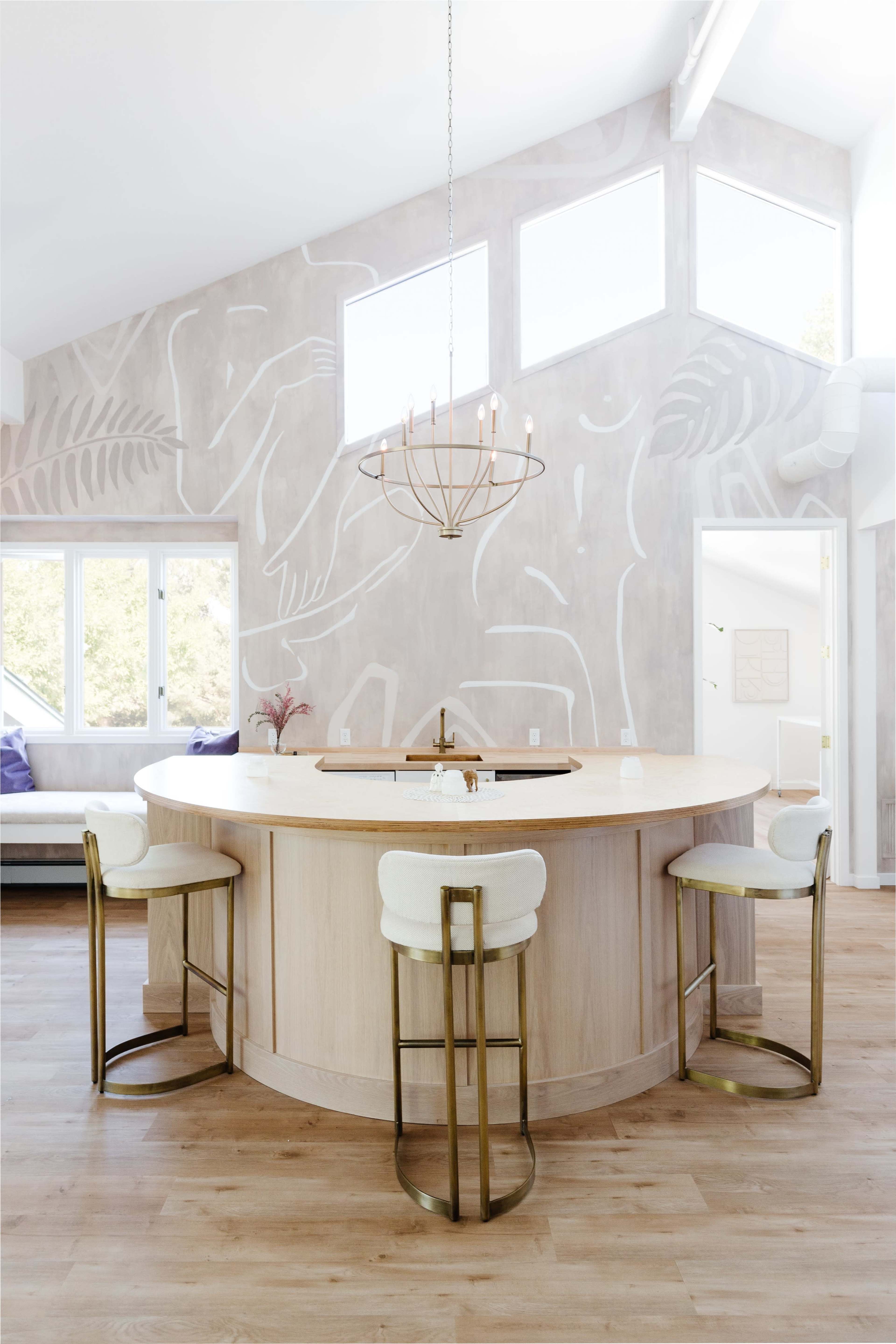 A circular wooden kitchen island with four upholstered bar stools and a modern chandelier, set against a backdrop of large windows and a light-colored mural on the wall.
