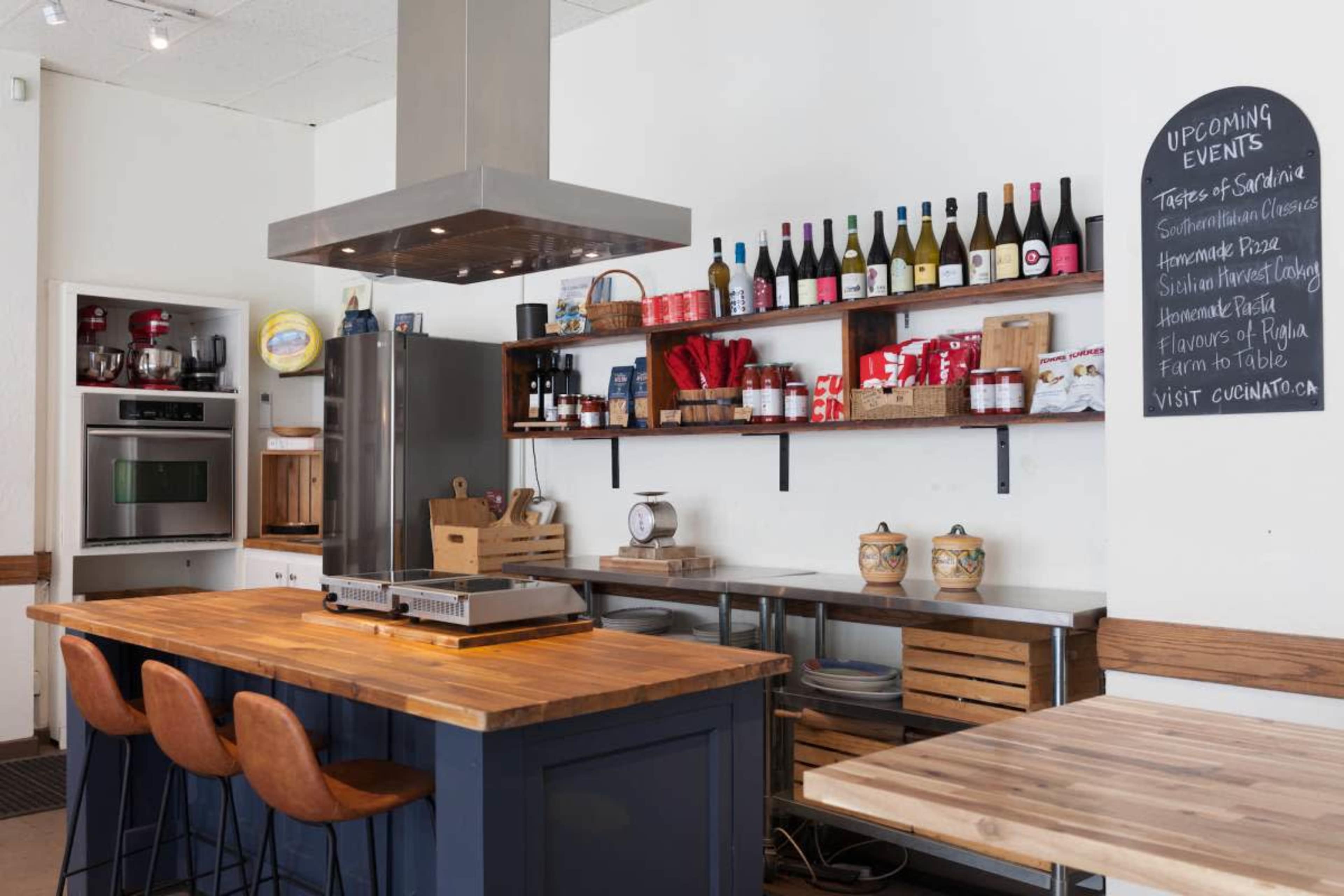 The image shows a modern kitchen with a large wooden island, several shelves stocked with kitchenware and bottles, and a chalkboard listing upcoming events.