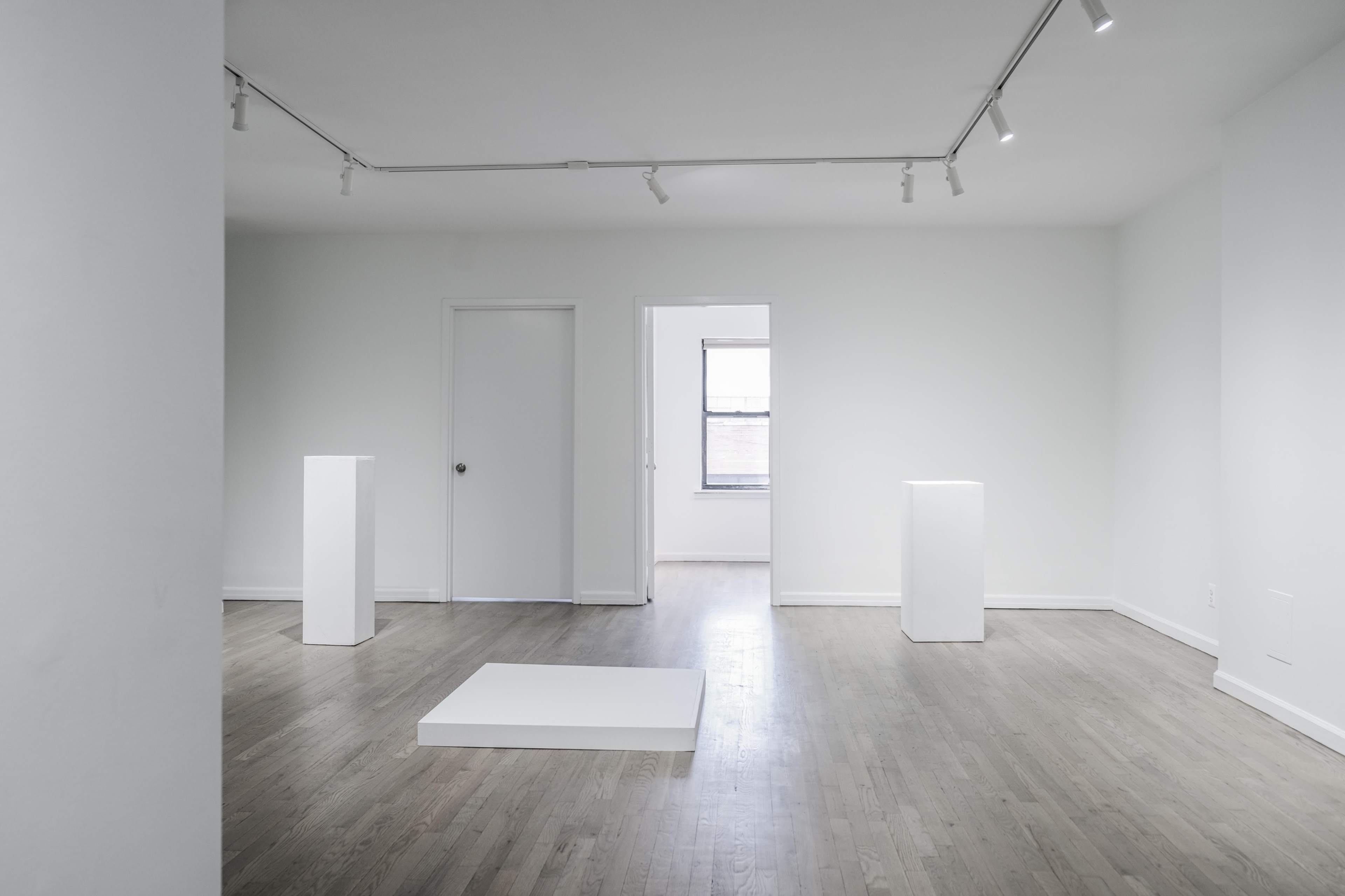 A minimalist art gallery with white walls, wooden floors, and three white pedestals arranged within the space.