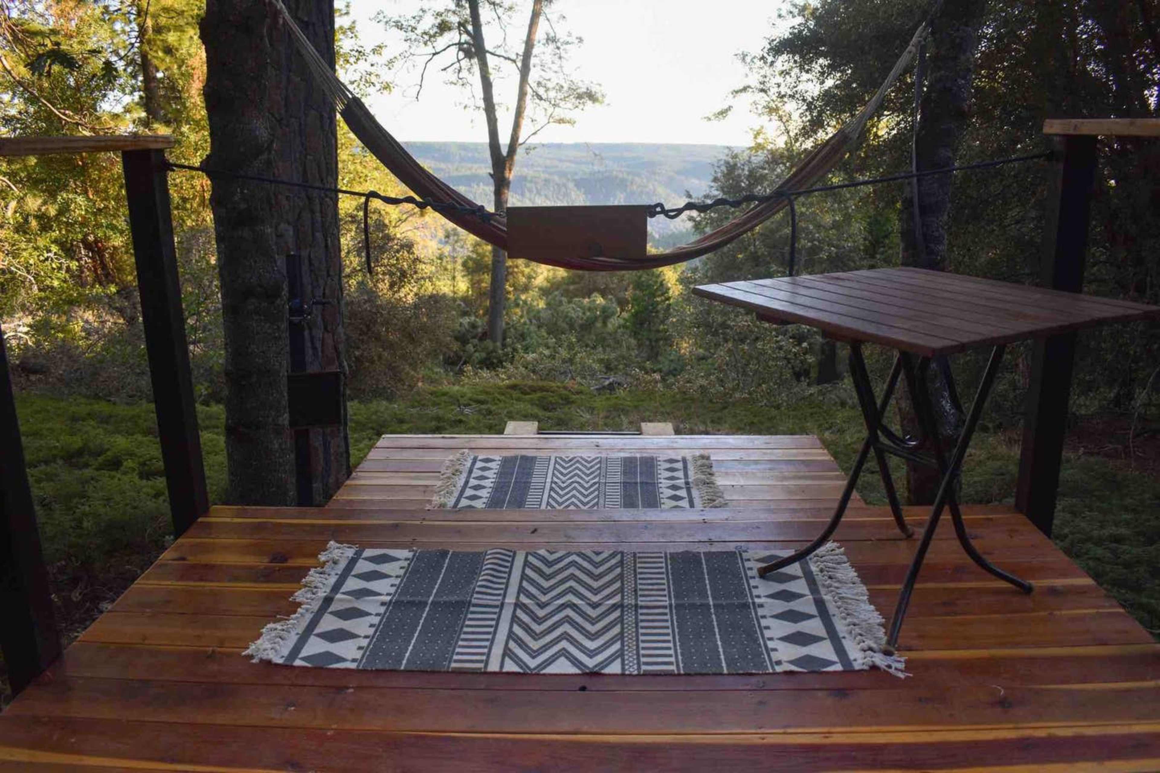 A wooden deck with a hammock suspended between two trees, accompanied by a small table and patterned rugs, overlooking a scenic view of hills and trees.