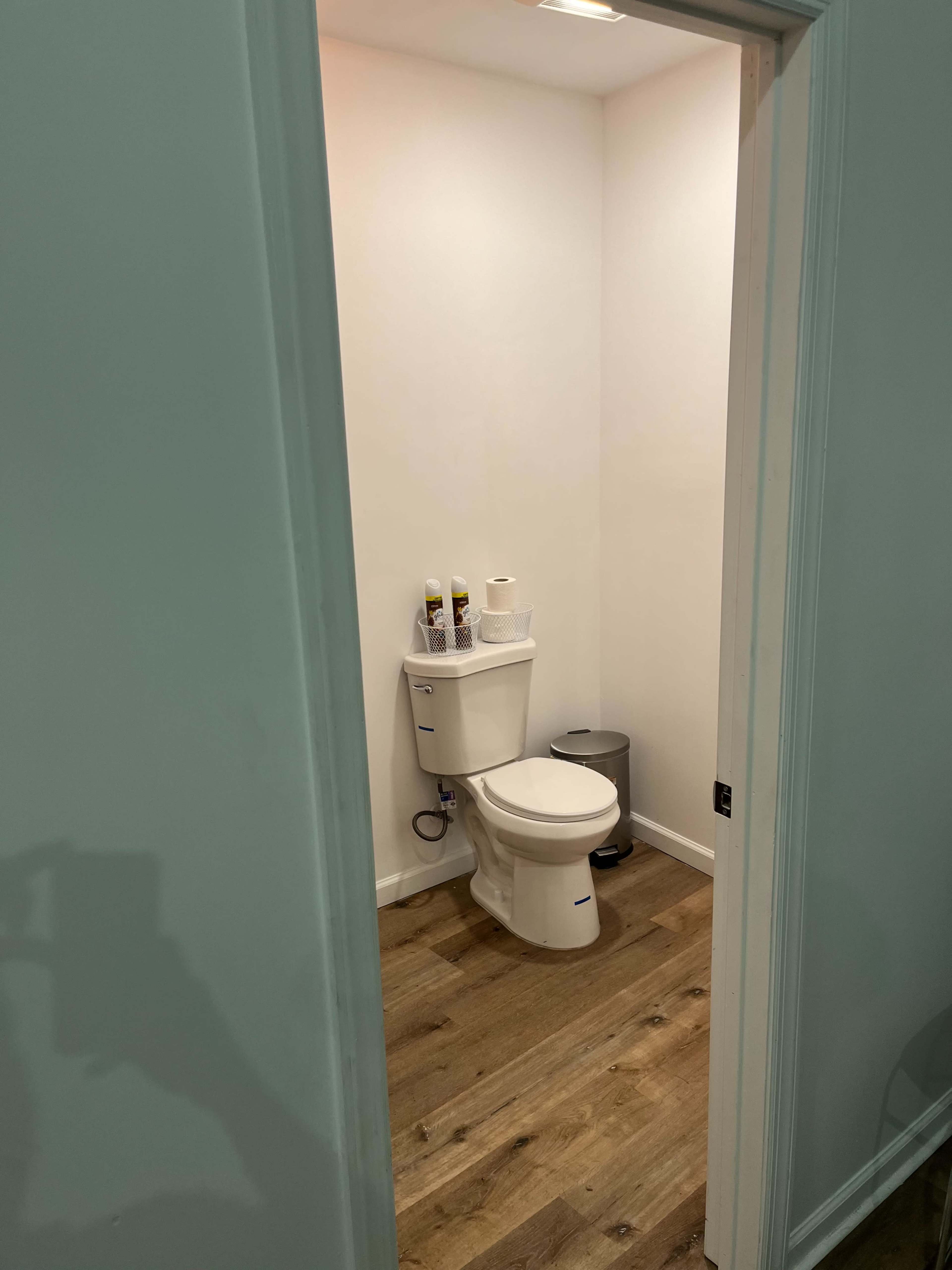 A small bathroom with a toilet, toilet paper, and a waste bin on a wooden floor.