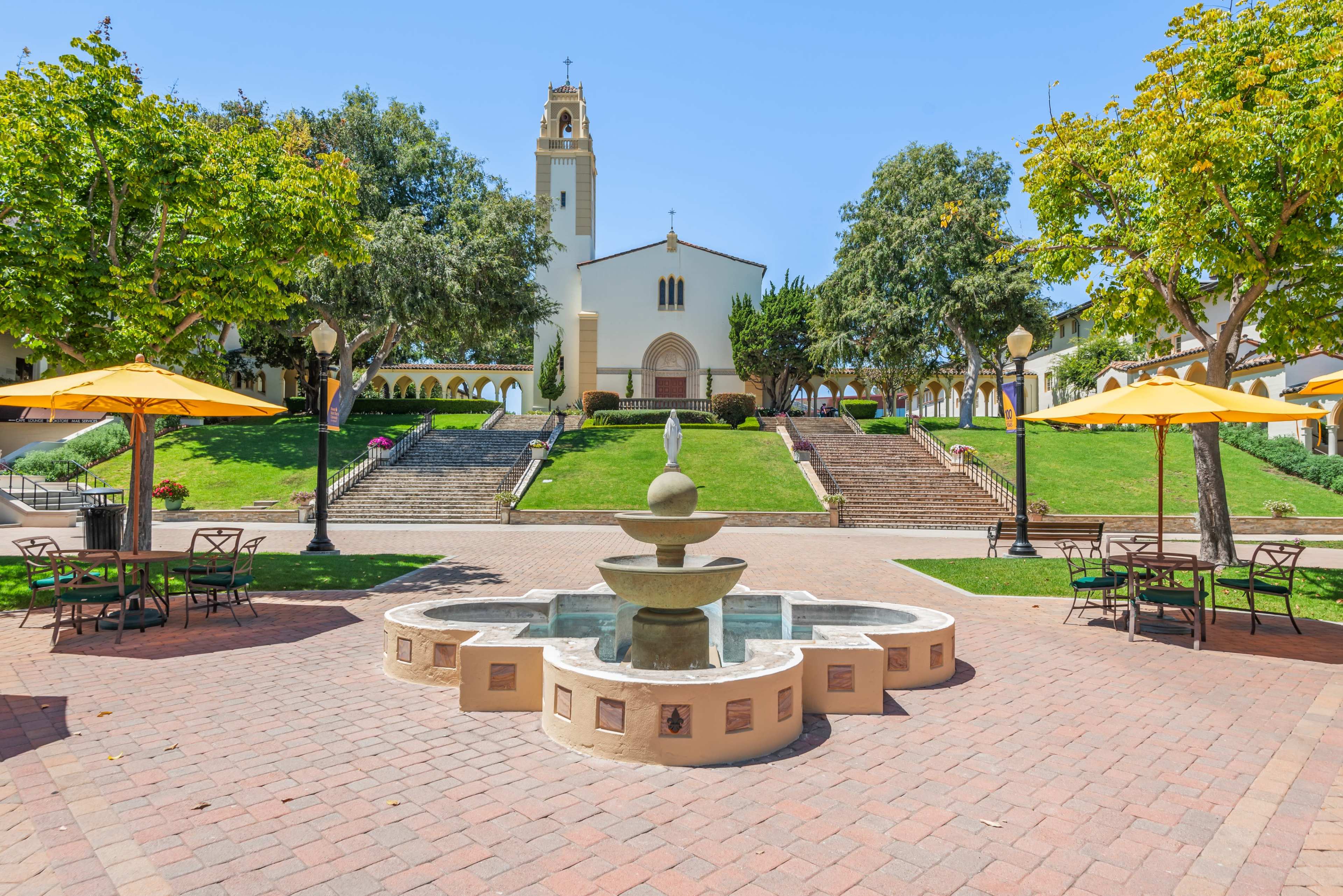 A fountain is centered in a plaza surrounded by tables and umbrellas, with a church and steps in the background.