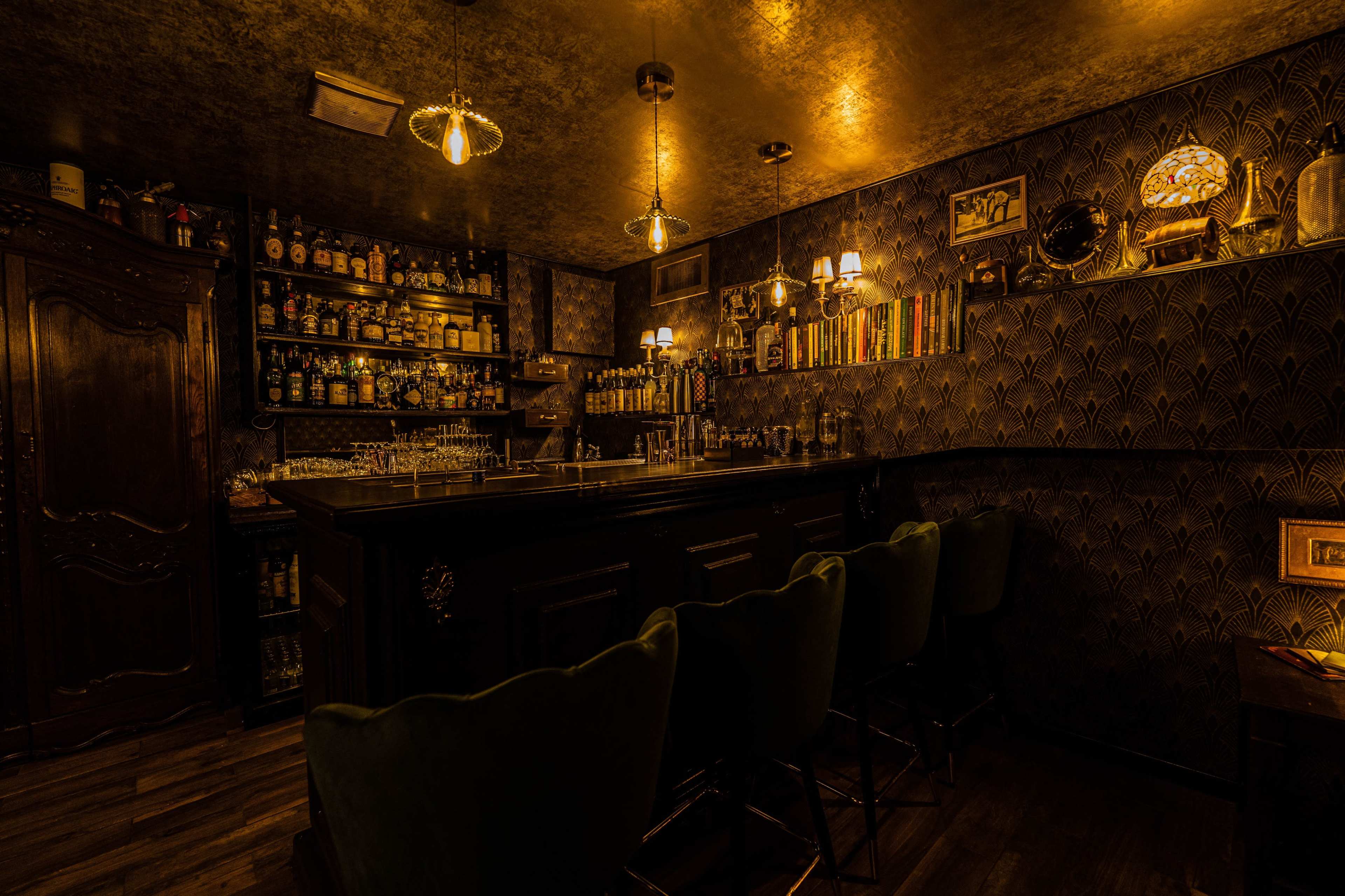 A dimly lit bar features a wooden counter with several chairs and shelves lined with an extensive array of liquor bottles.