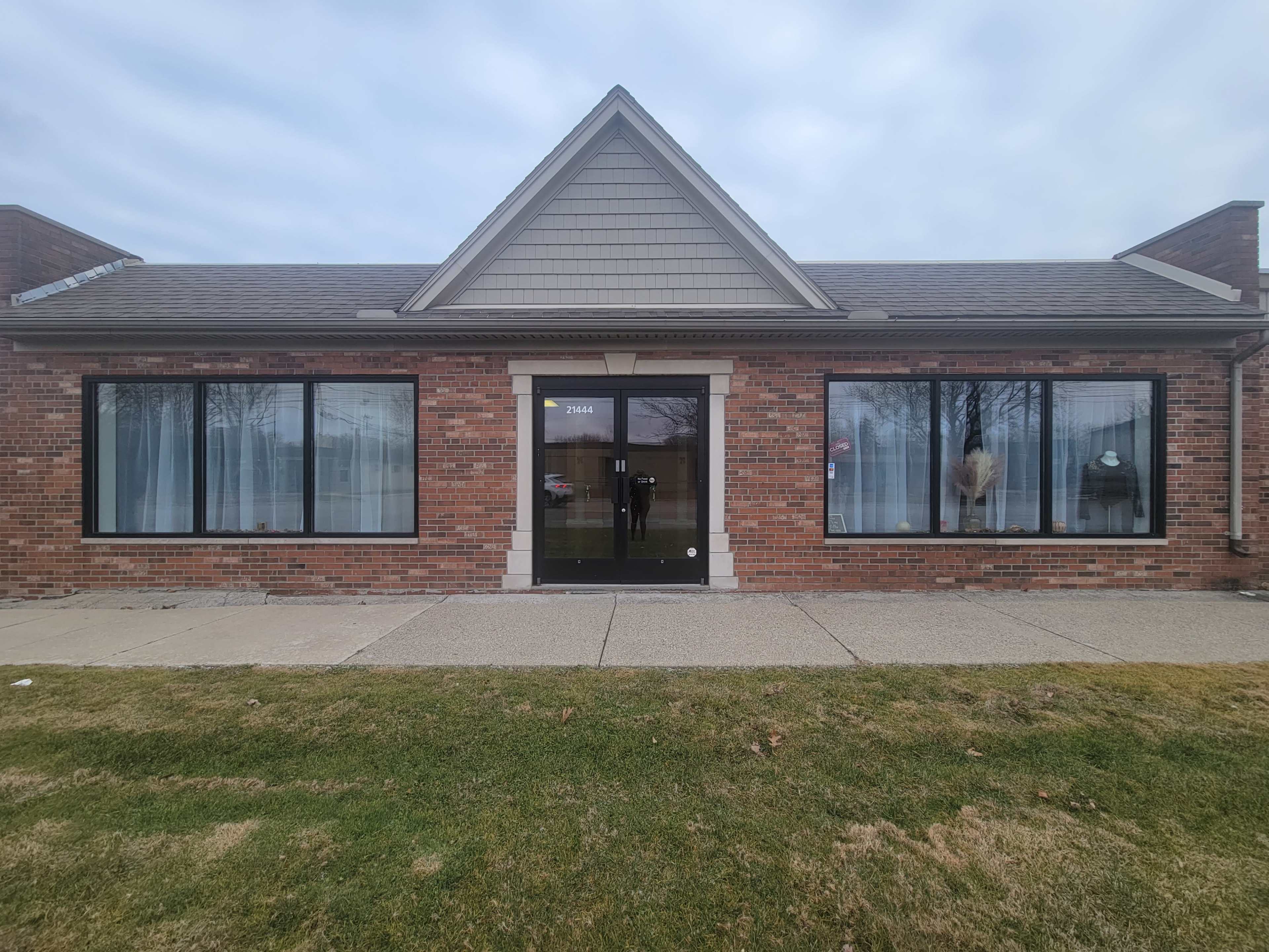 The image shows a brick building with large windows and a front door, set against a grassy area and cloudy sky.