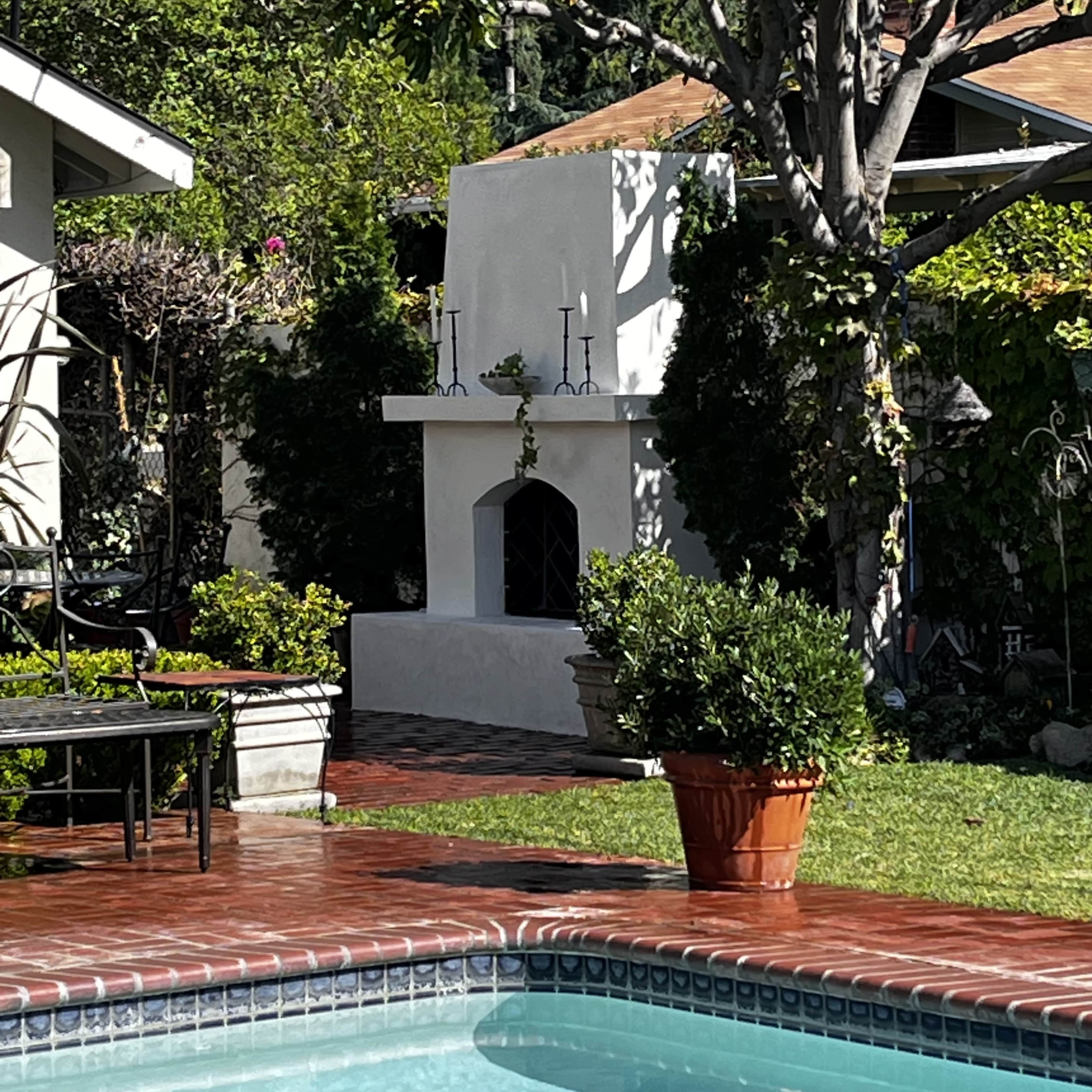 The image shows a poolside area featuring a brick patio, potted plants, and a white outdoor fireplace surrounded by greenery.