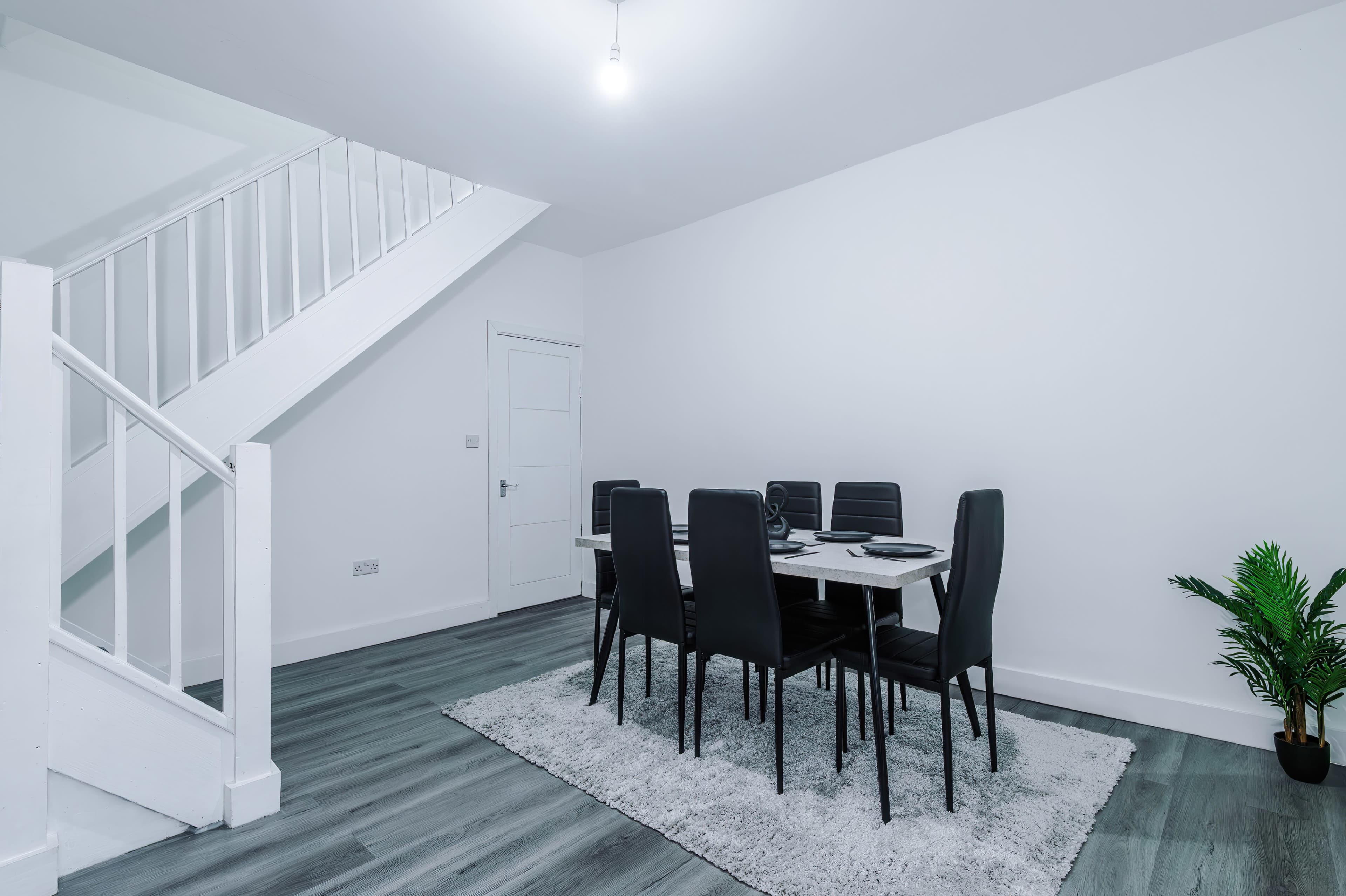 A dining area features a table with chairs on a rug, set beneath a staircase in a brightly lit room.