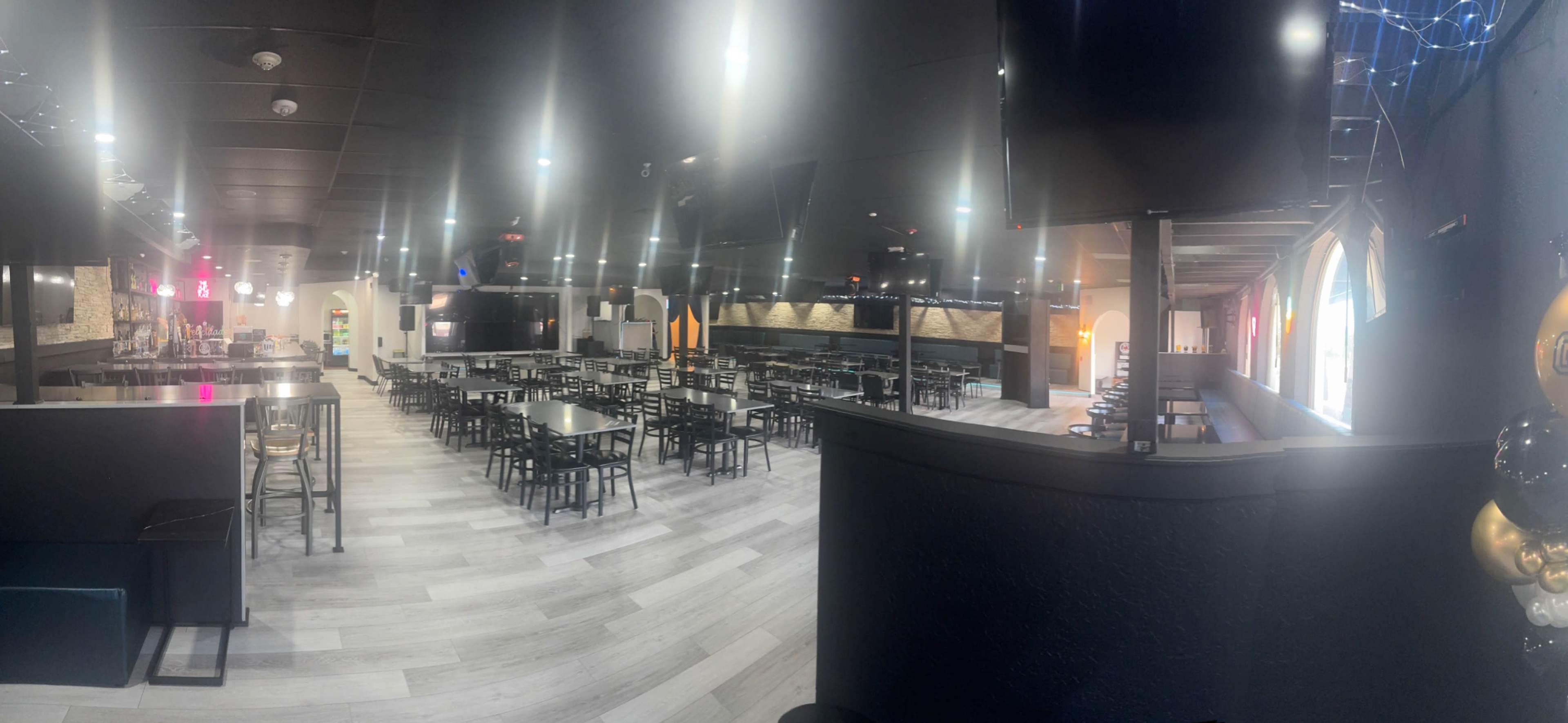 The image shows a large, modern restaurant interior with multiple black tables and chairs arranged under bright overhead lights.