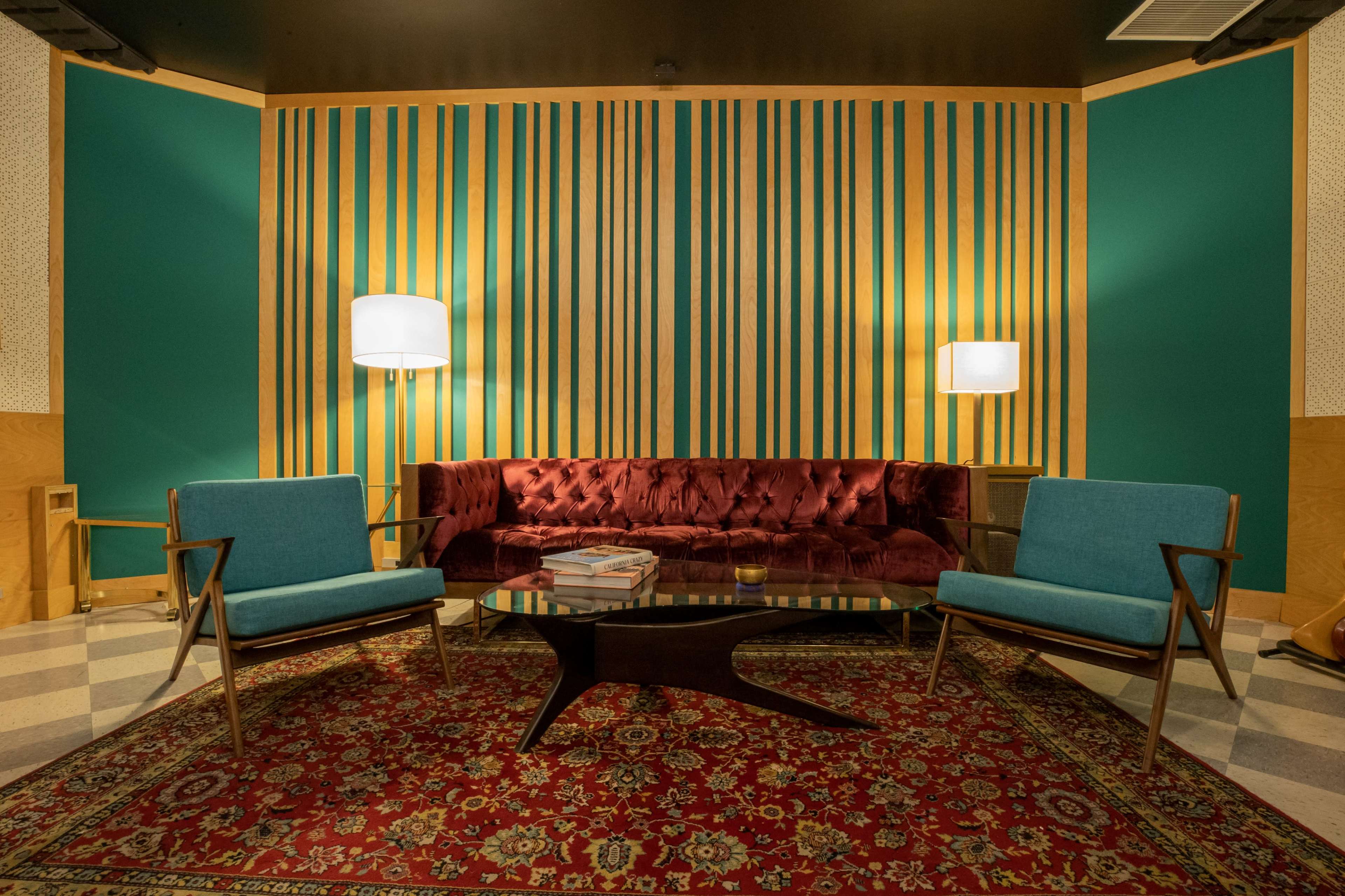 The scene features a living area with a red velvet sofa, two teal chairs, a circular glass coffee table, and a patterned area rug against a backdrop of striped green and wood-paneled walls.