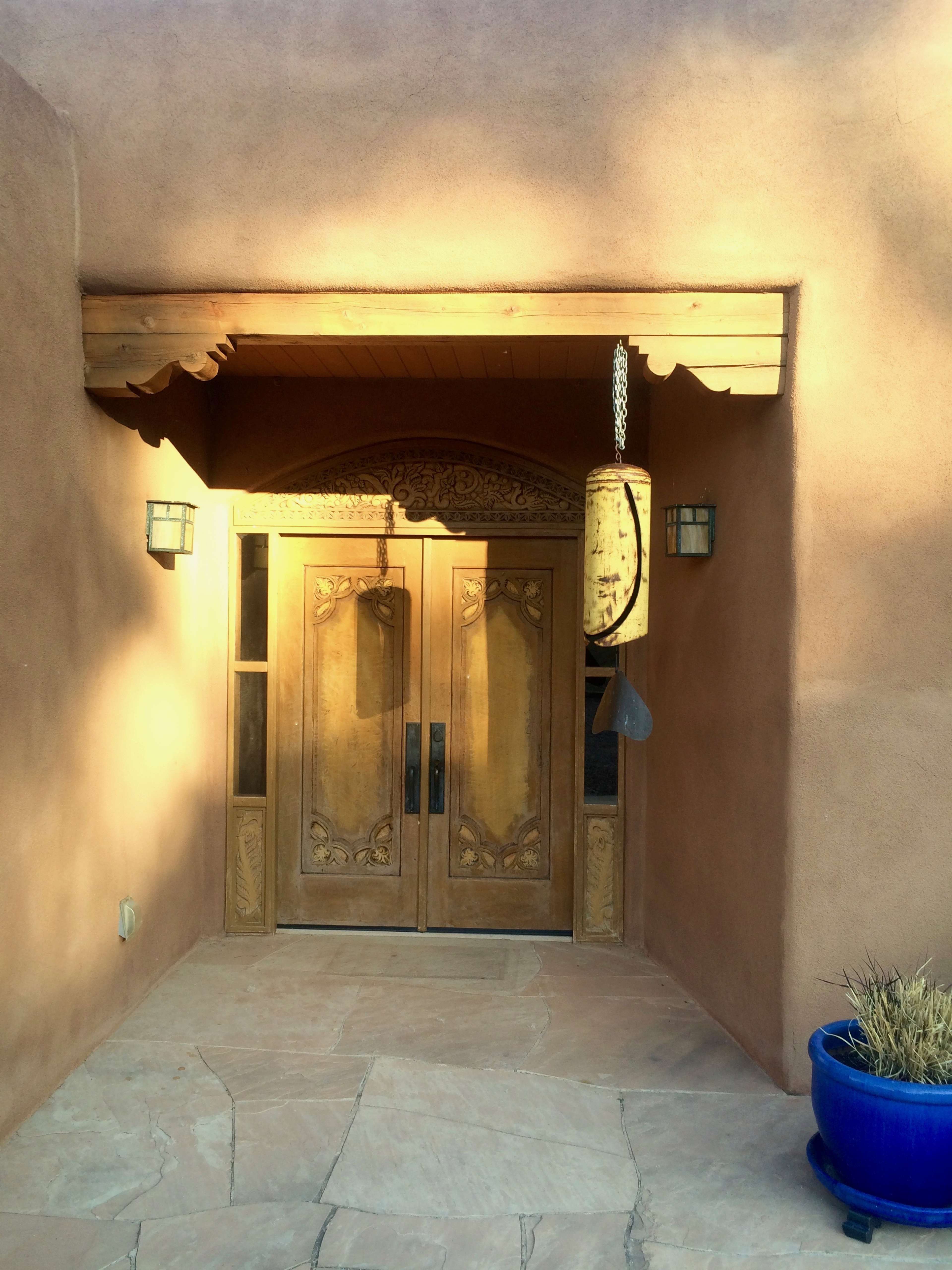 A decorative wooden double door under a covered entryway, framed by adobe walls and accompanied by a blue planter on the right.