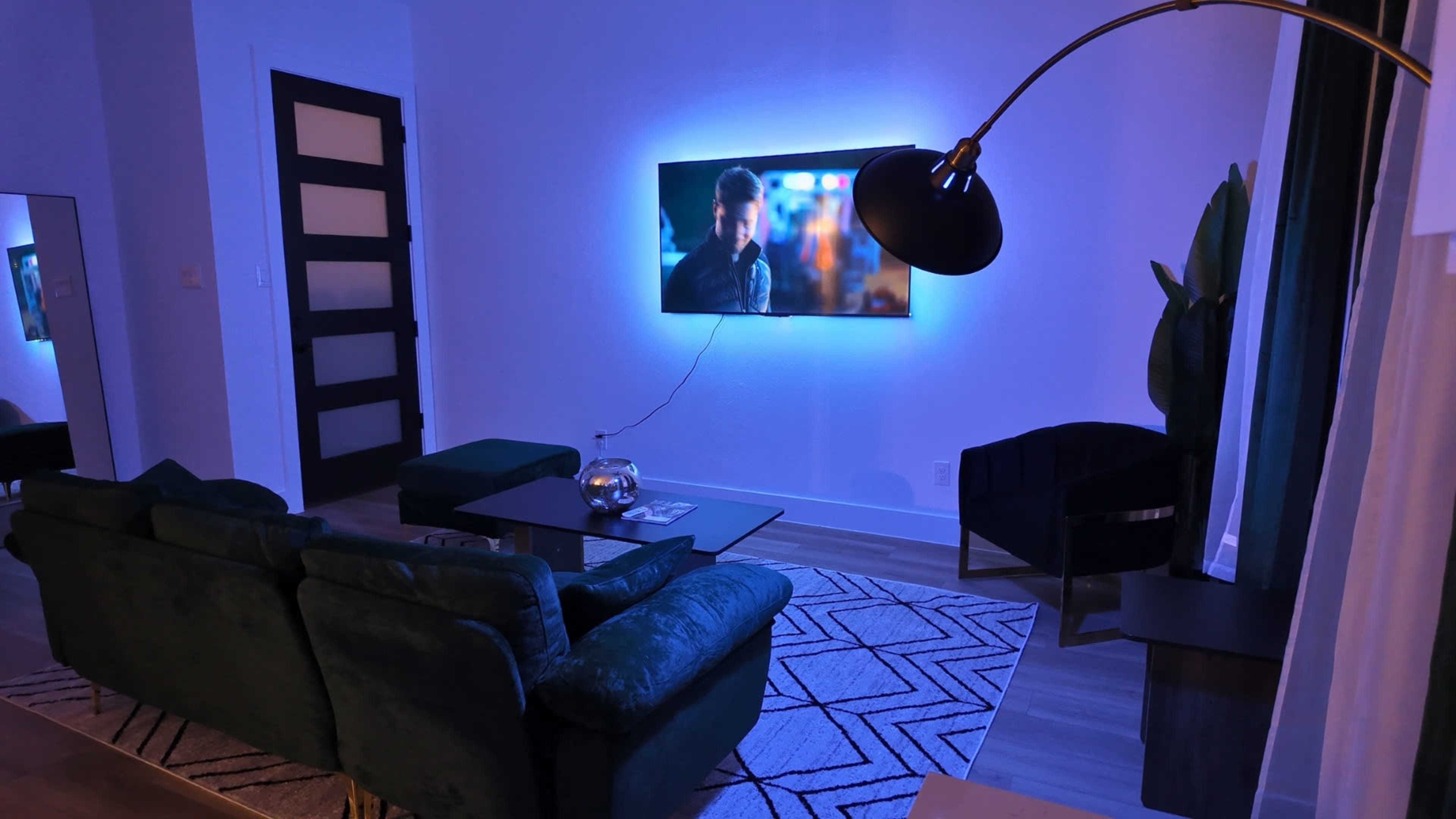 The living room features a sofa and a coffee table, with a television mounted on the wall, illuminated by blue lighting.