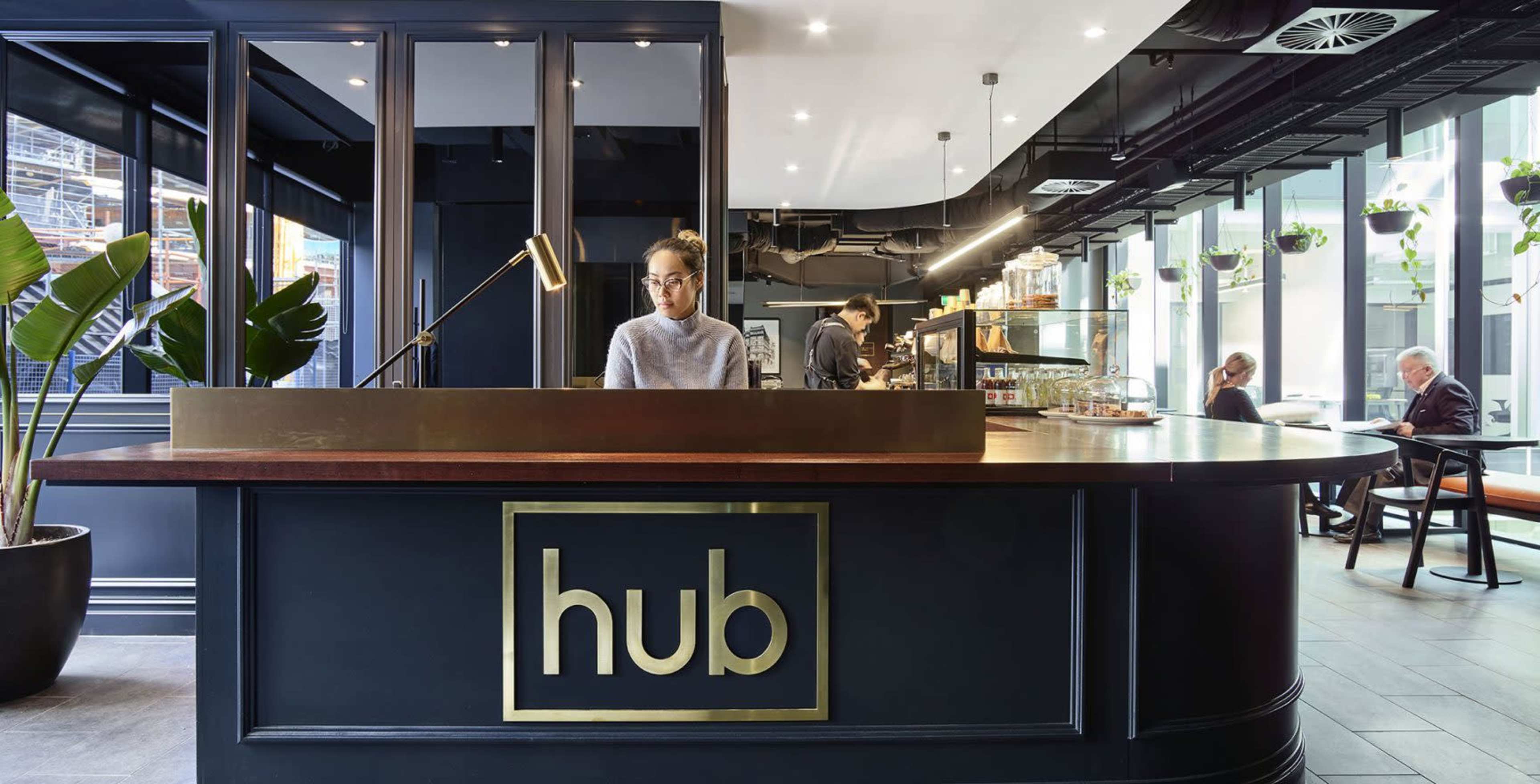 A modern reception area features a dark counter labeled "hub," with a desk attendant and a cafe space in the background.