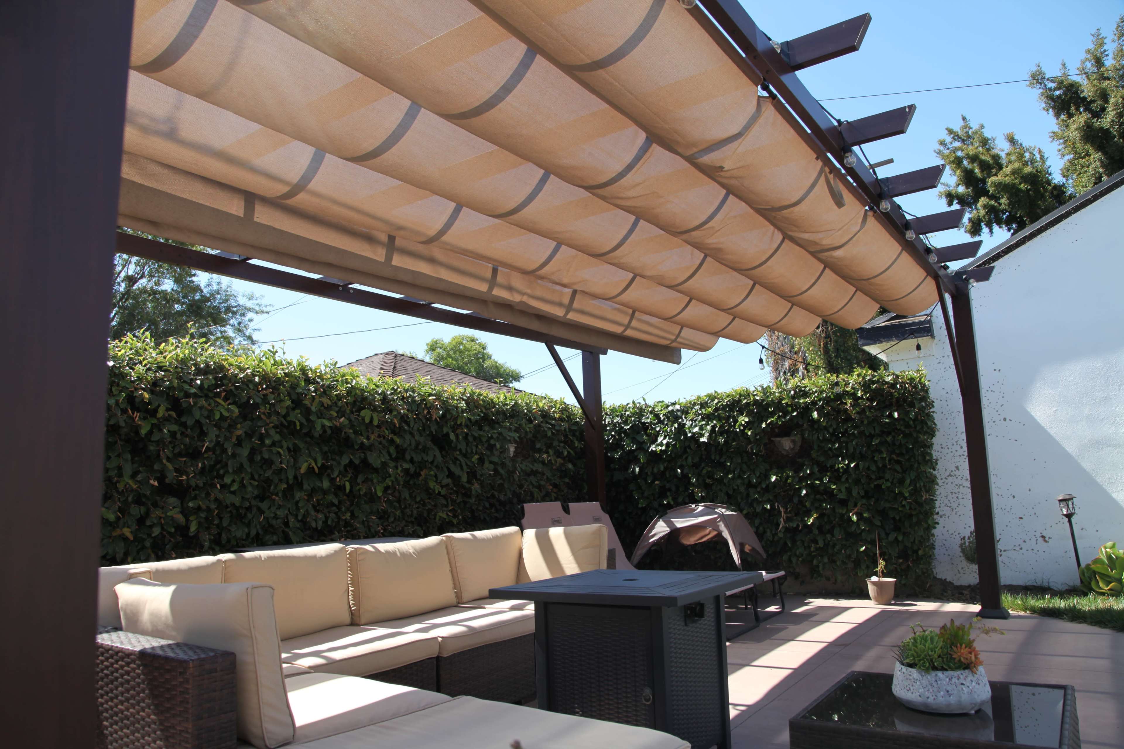 A wooden pergola with a retractable canopy covers a seating area featuring beige cushions and a dark table, surrounded by a green hedgerow.