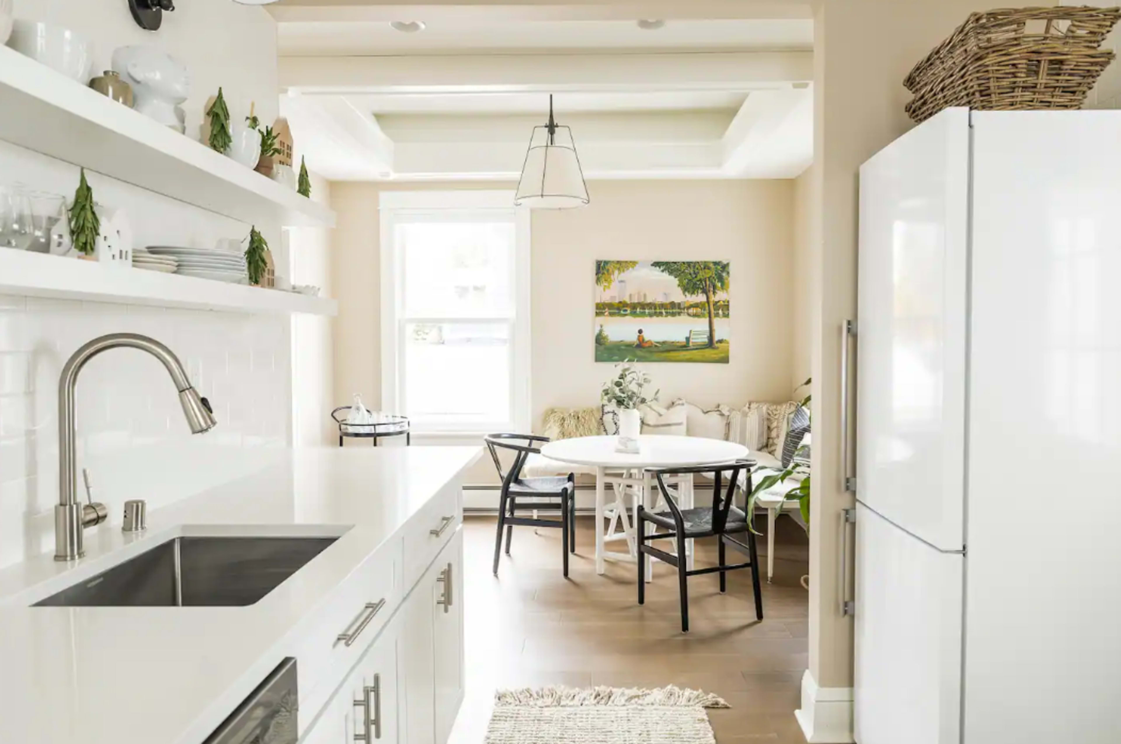 A bright kitchen features white cabinetry and a sleek sink, leading into a dining area with a round table and a colorful painting on the wall.