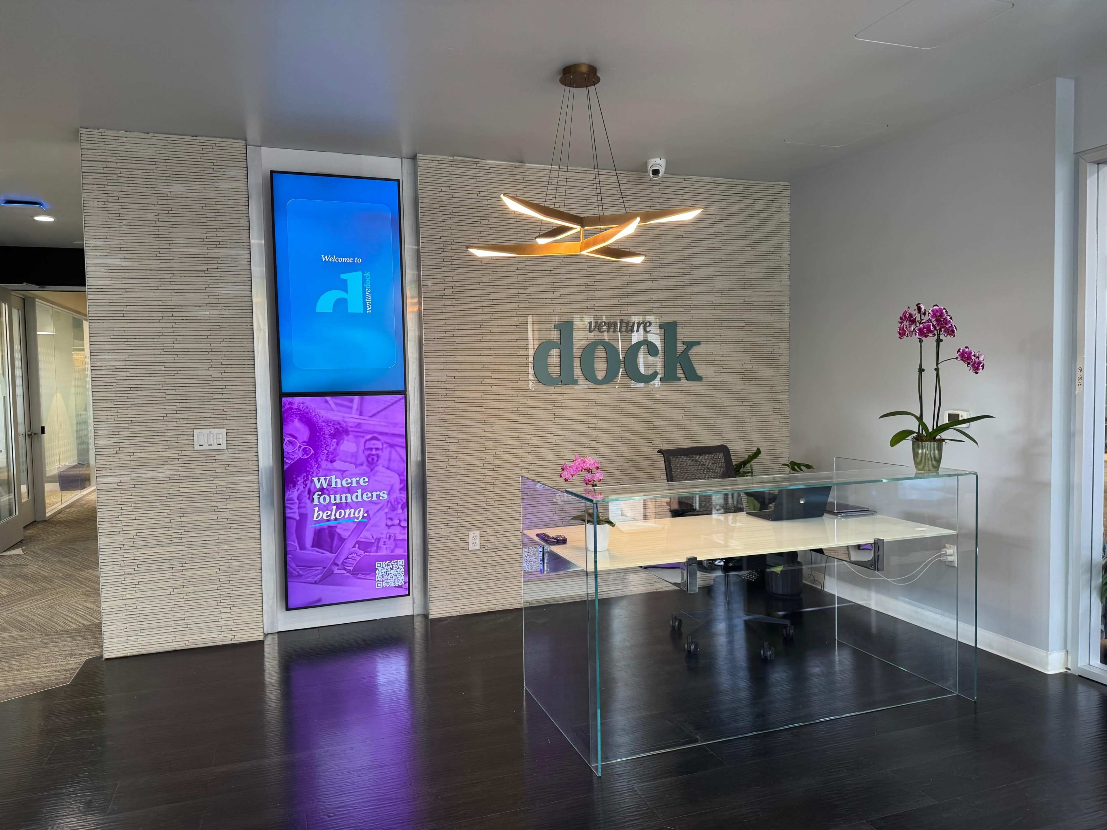 The image shows a modern reception area with a clear glass front desk, a large wall sign reading "dock," and digital screens displaying welcoming messages and graphics.