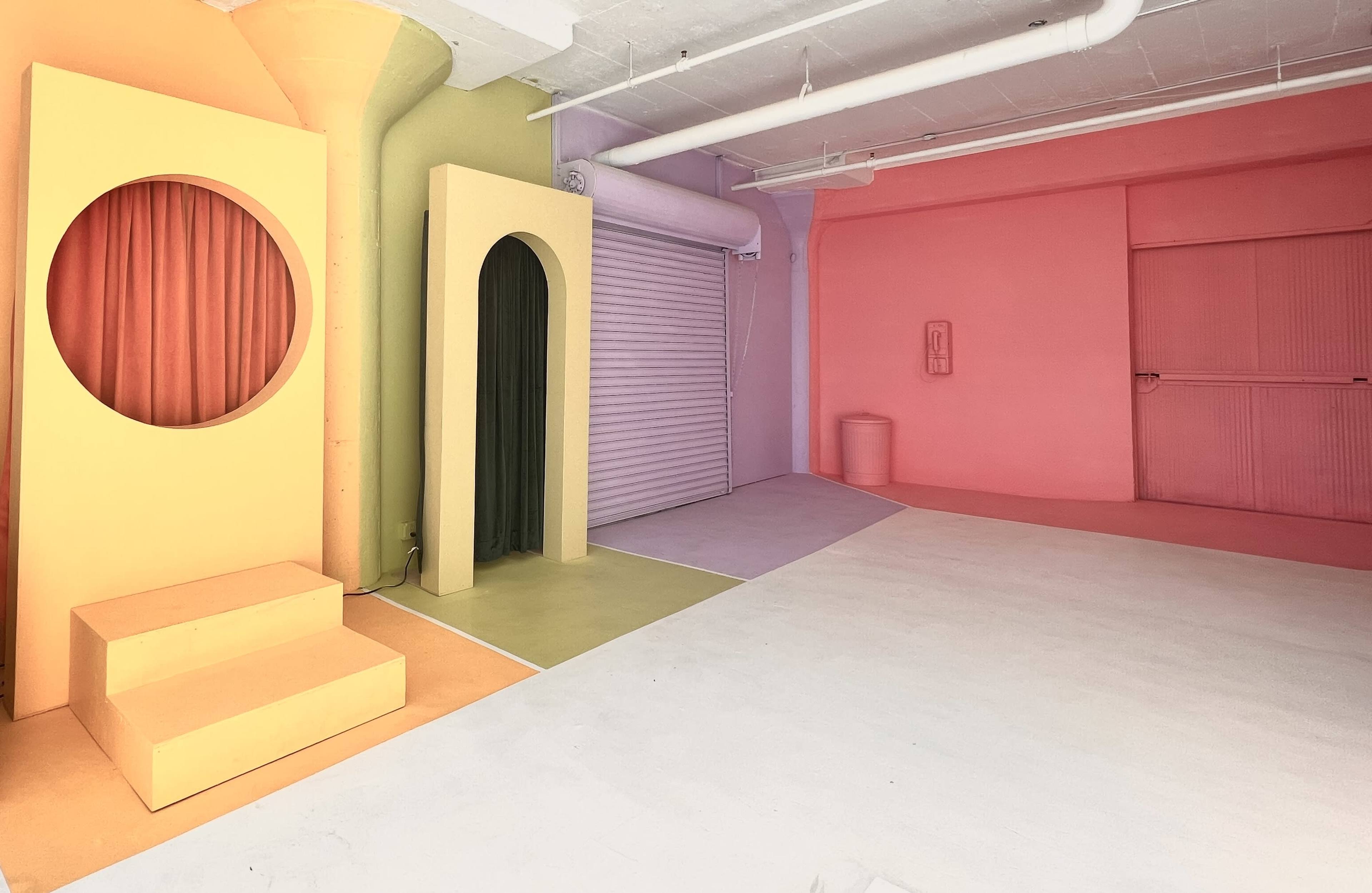 The scene features a brightly colored studio space with geometric shapes, including a yellow structure with steps and circular openings, and walls in pastel shades of pink, green, and orange.