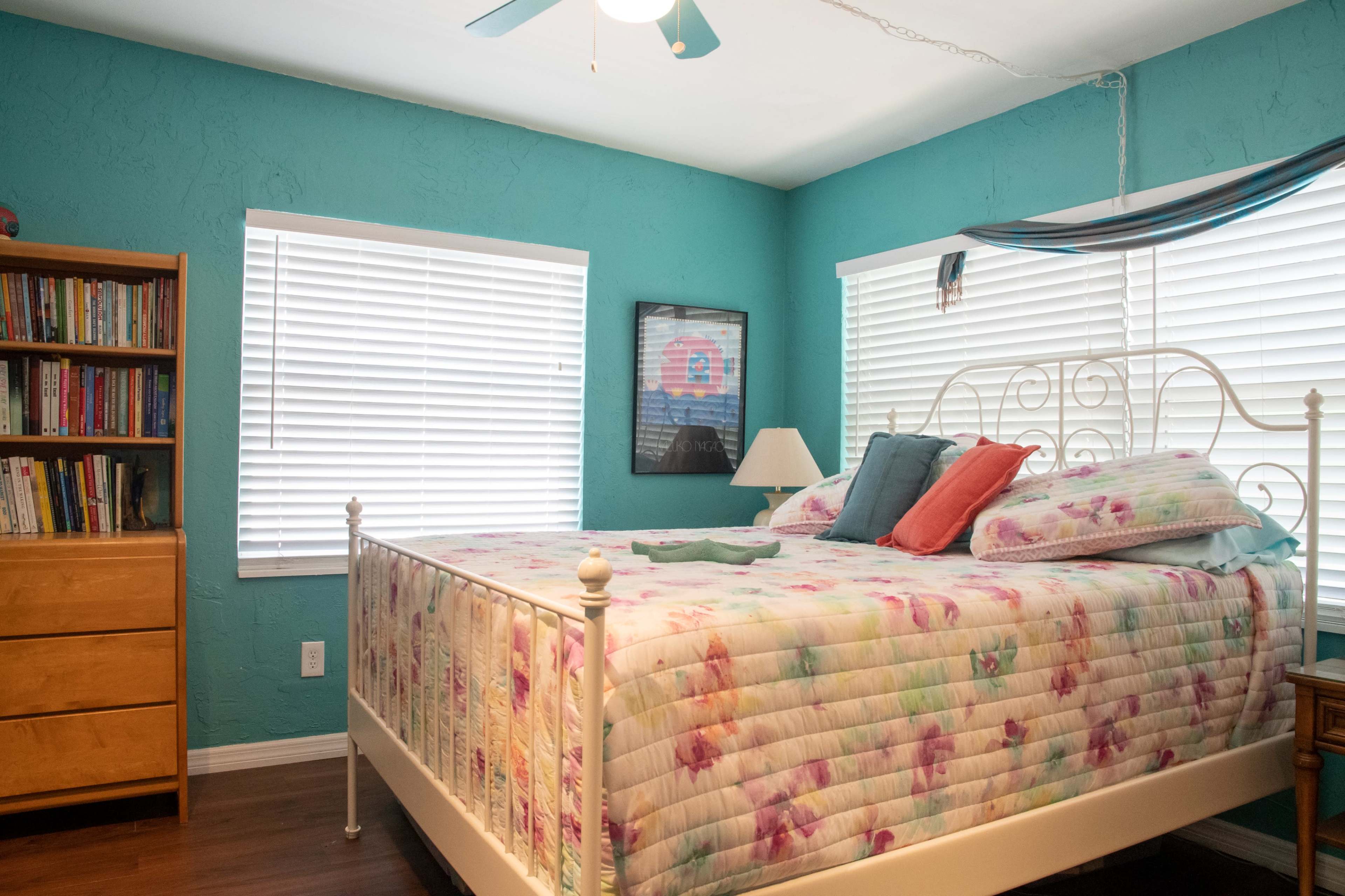 A brightly colored bedroom features a bed with a floral quilt, two accent pillows, a wooden bookshelf, and a chest of drawers.
