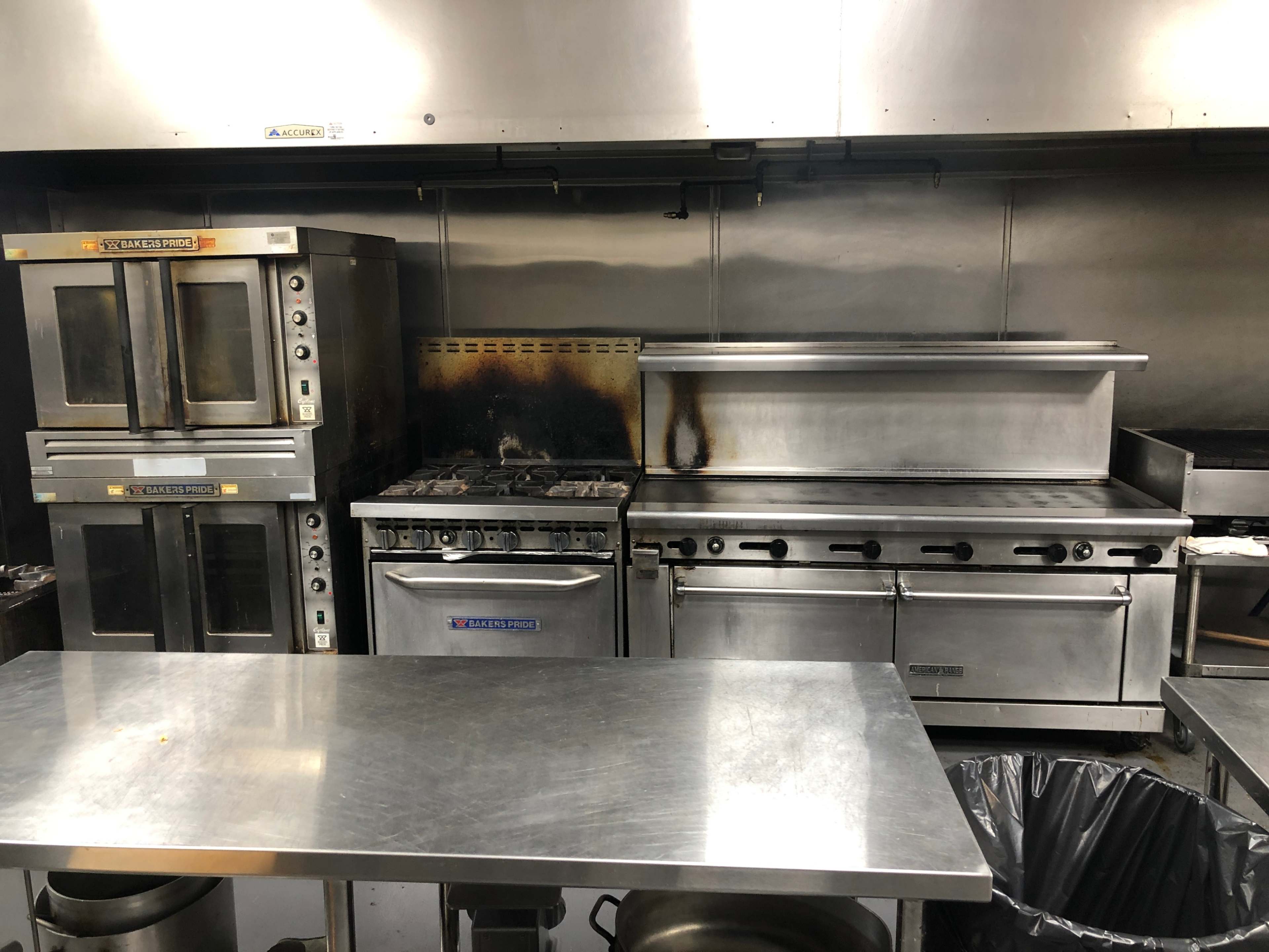 The image shows a stainless steel commercial kitchen featuring an oven, a stove, and a grill, with a metal table and a trash bin in the foreground.