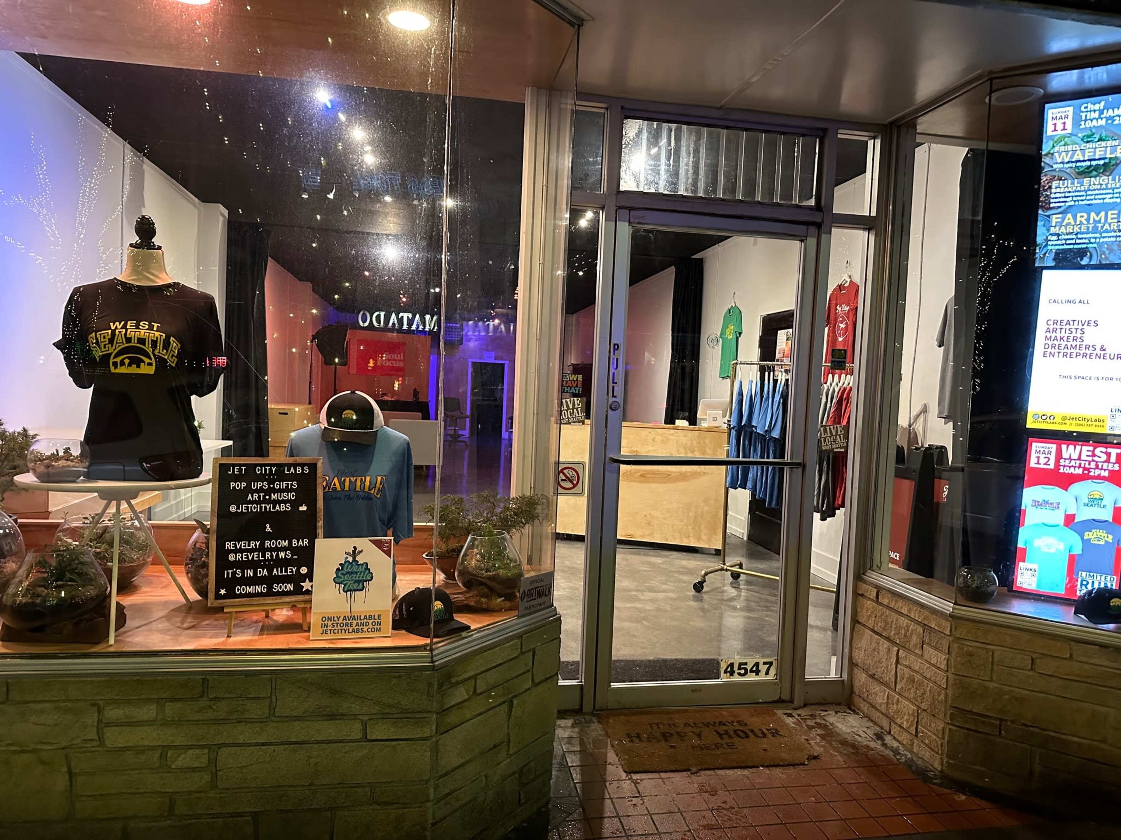 The storefront displays t-shirts and promotional signs in a well-lit window, showcasing a variety of merchandise and upcoming events.