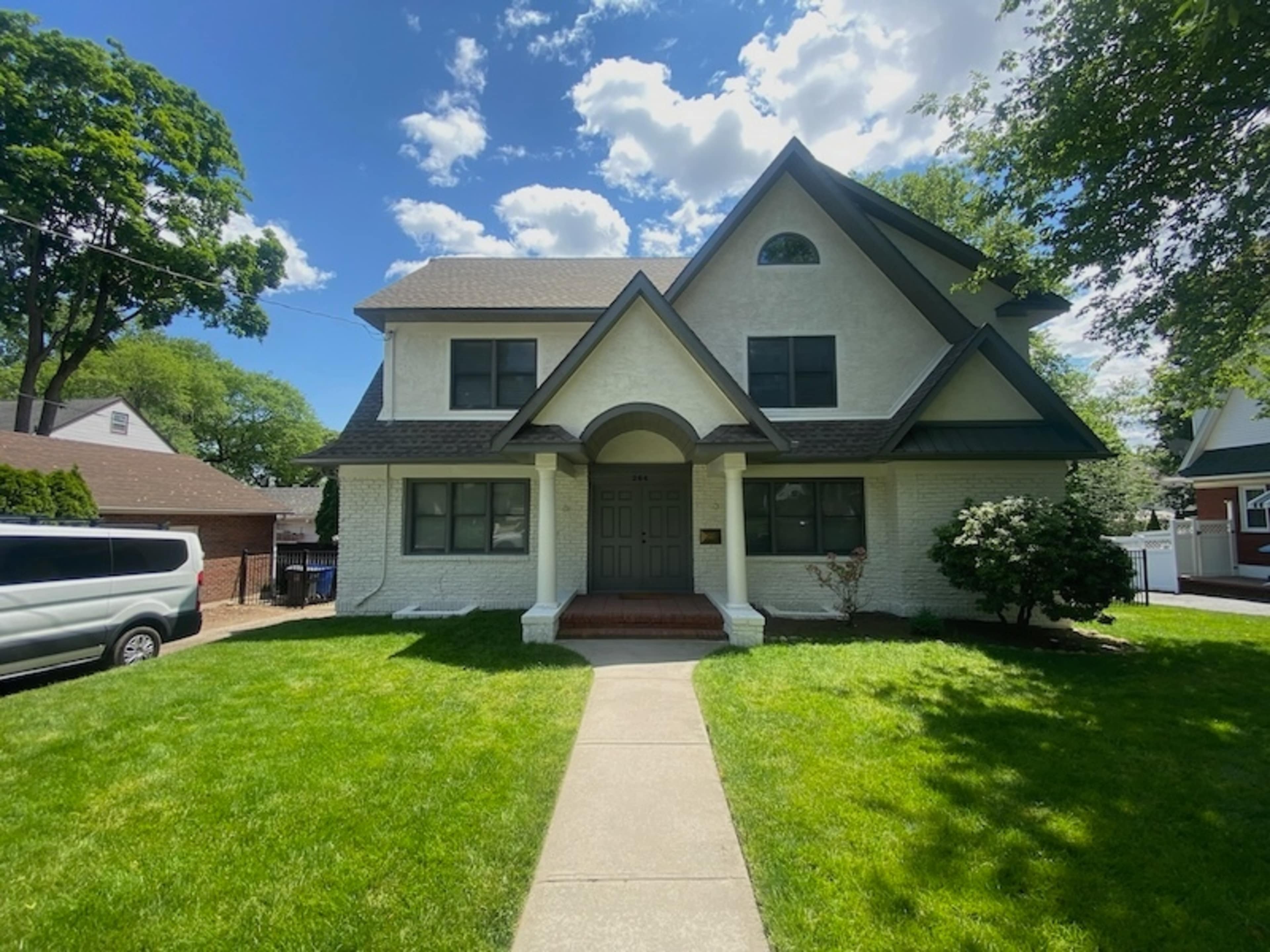 A two-story white house with a peaked roof, large windows, and a front walkway leading to a covered entrance, surrounded by grass and trees.