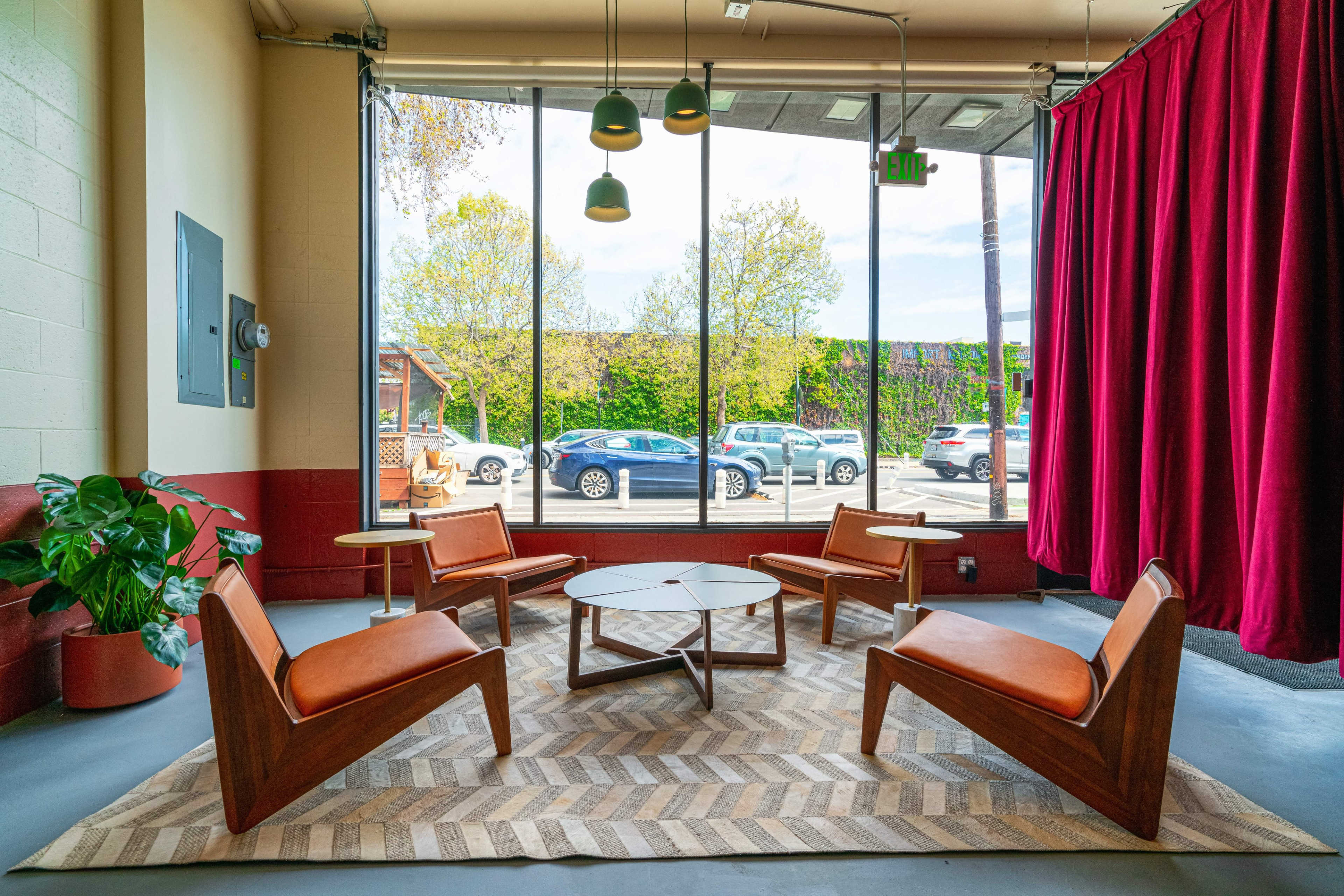 A cozy seating area features four wooden chairs with orange cushions arranged around a circular table on a patterned rug, set against a large window with natural light and a view of parked cars outside.