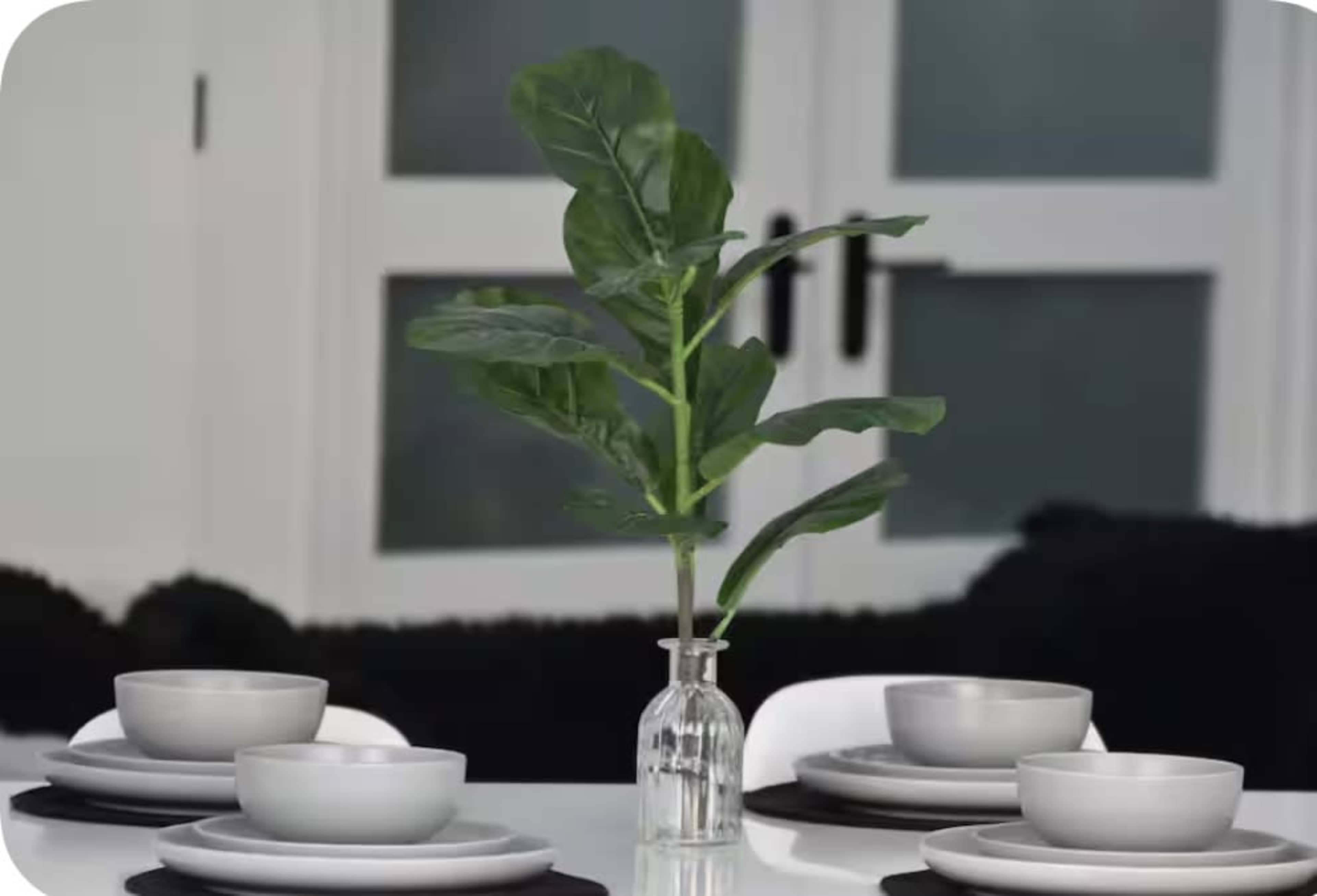 A dining table is set with gray plates and bowls, featuring a small glass vase containing a leafy green plant in the center.