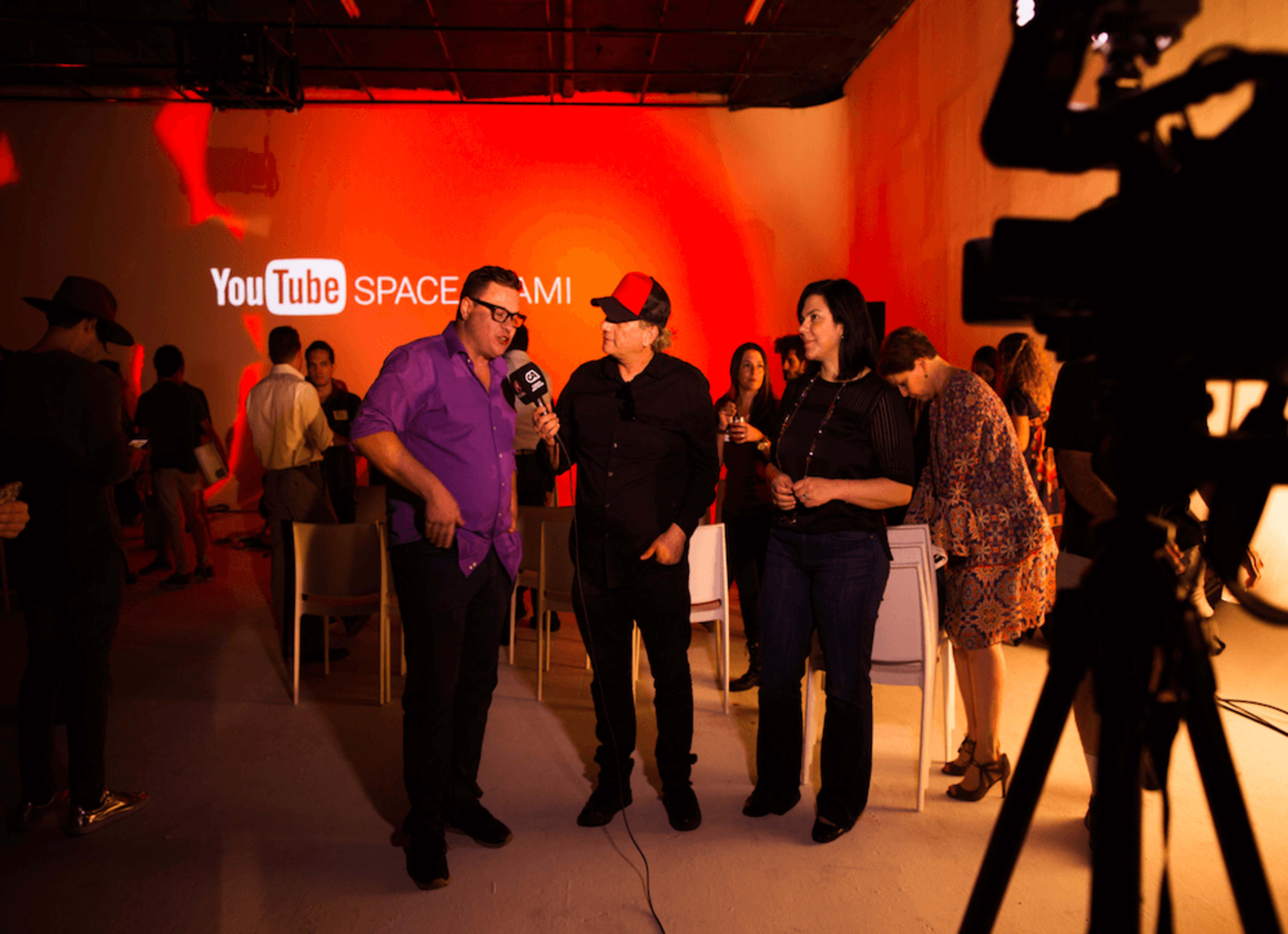 A group of people gathers in a brightly lit room with a "YouTube Space Miami" backdrop while two individuals conduct an interview in the foreground.
