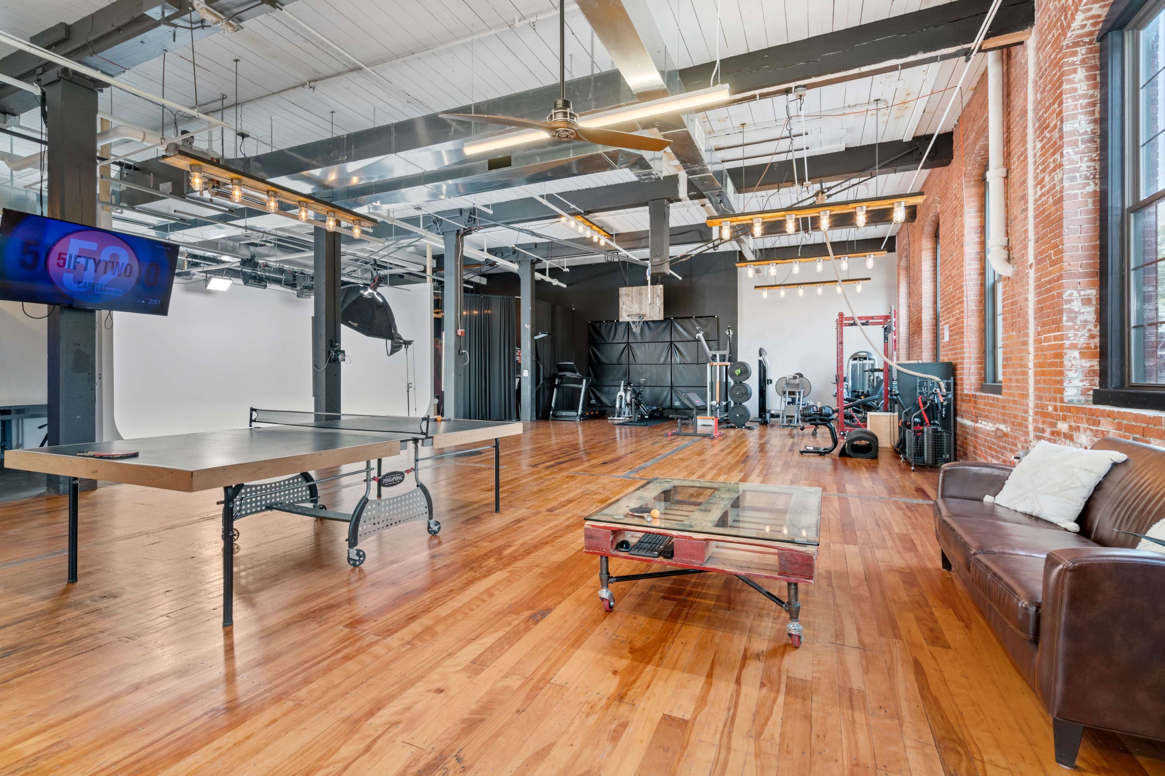 A spacious industrial-style studio featuring polished wooden floors, exposed brick walls, and various workout equipment arranged in a well-lit area.
