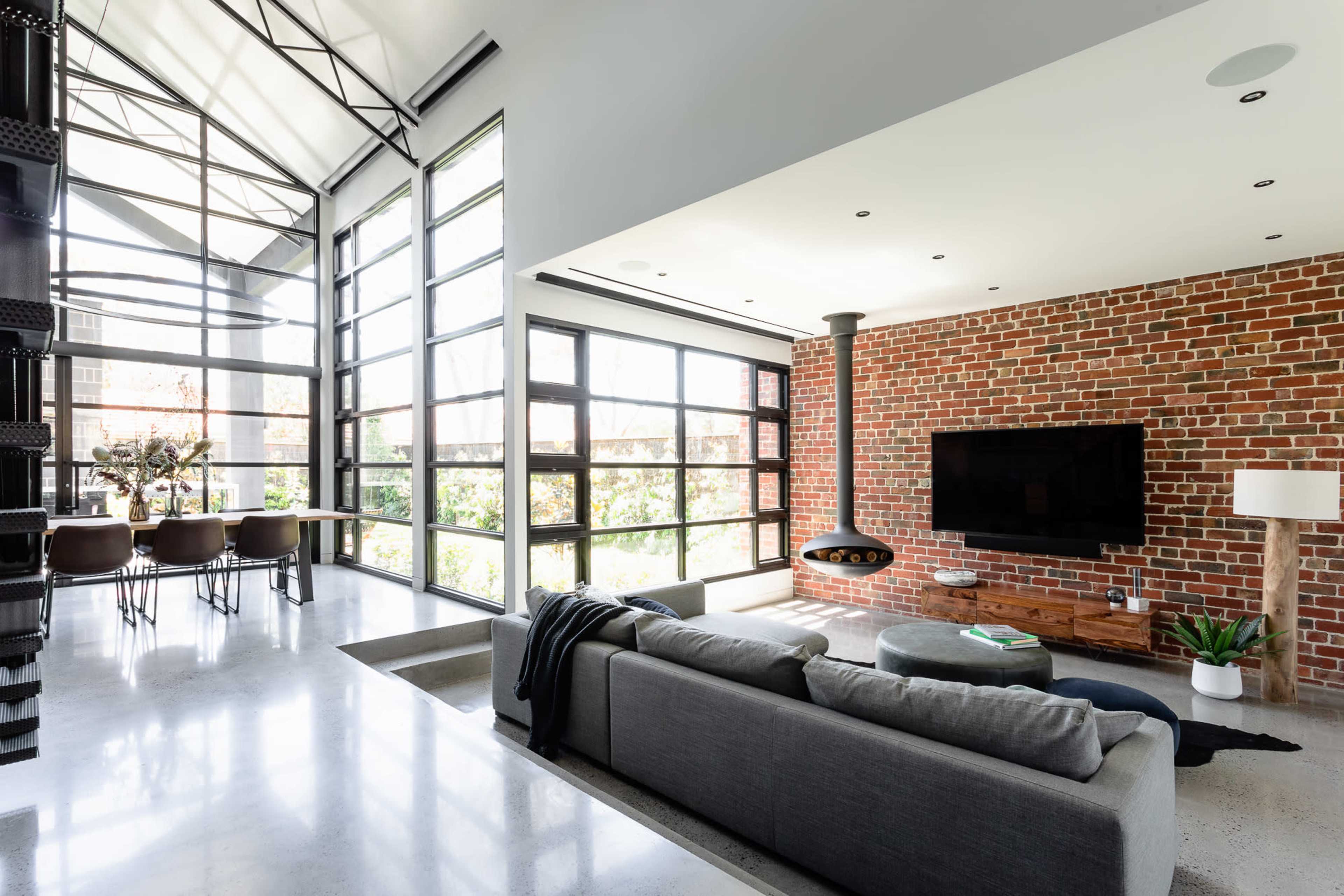 A modern living room features a gray sofa, a large brick wall with a mounted TV, and large windows allowing natural light to fill the space.