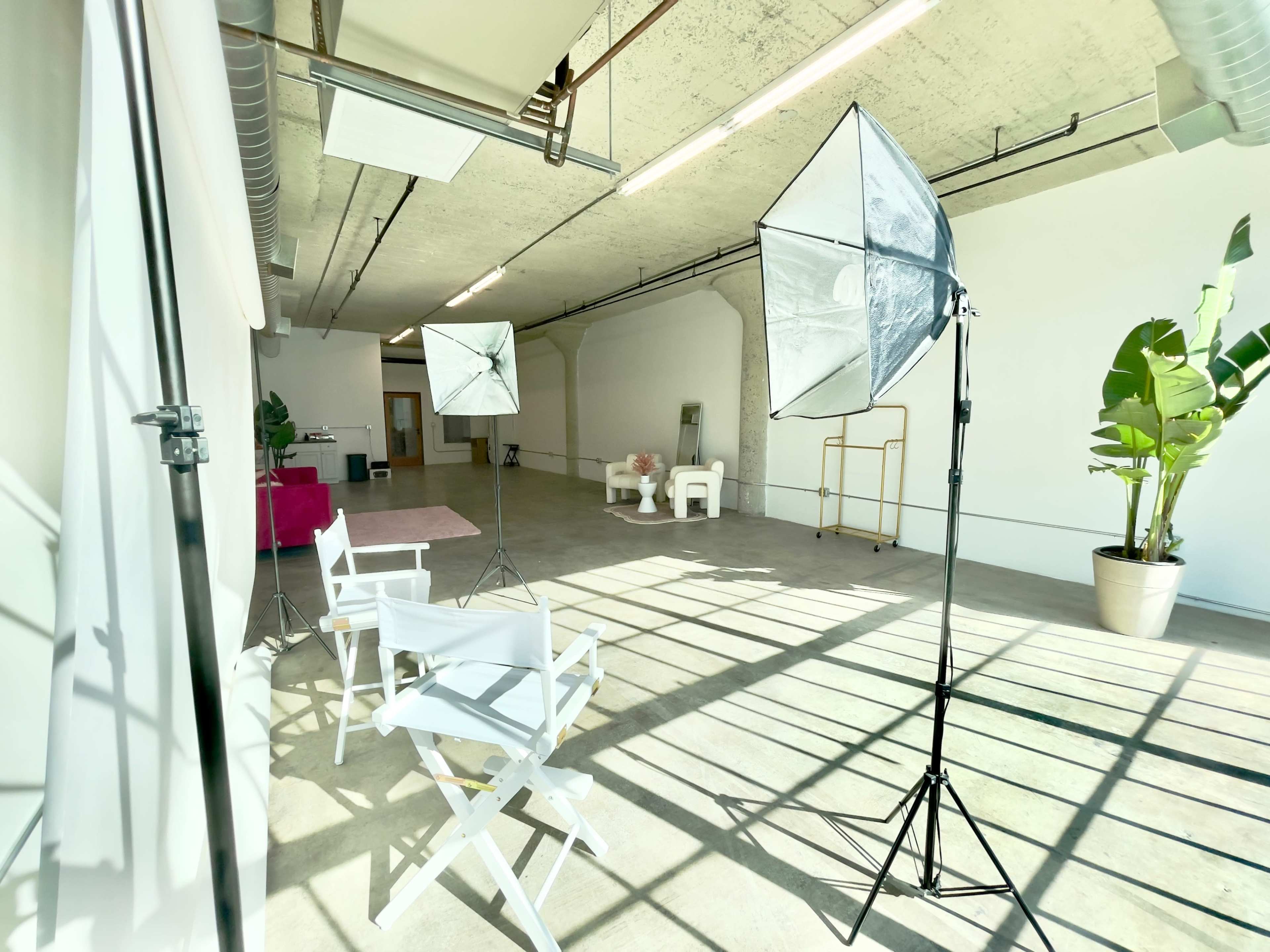 The image shows a spacious studio with concrete floors, a white backdrop, two light stands with softboxes, and two white director's chairs.