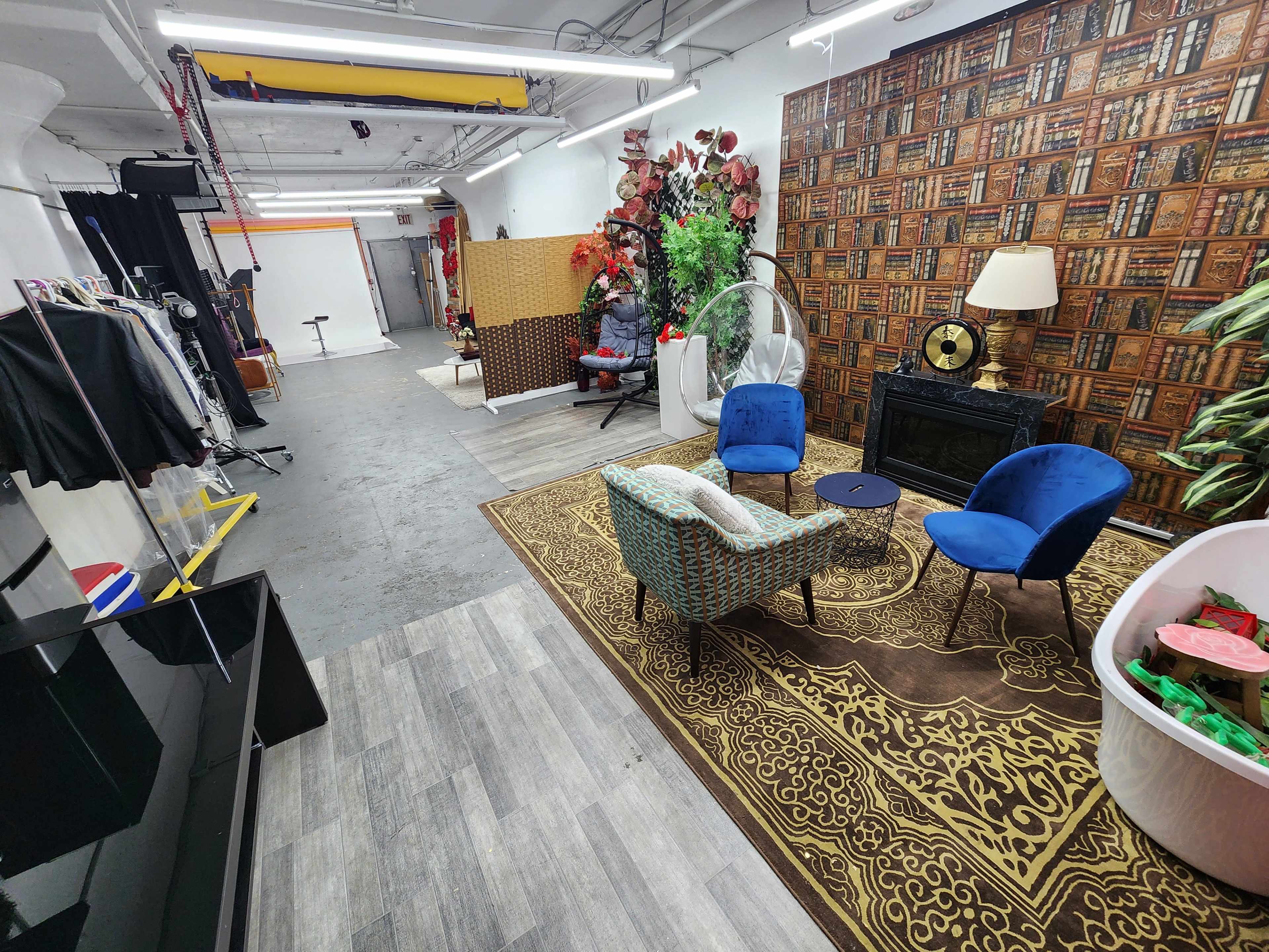 The image shows a spacious studio with a patterned wall, two blue chairs, a small table, decorative plants, and a variety of props and clothing along one side.