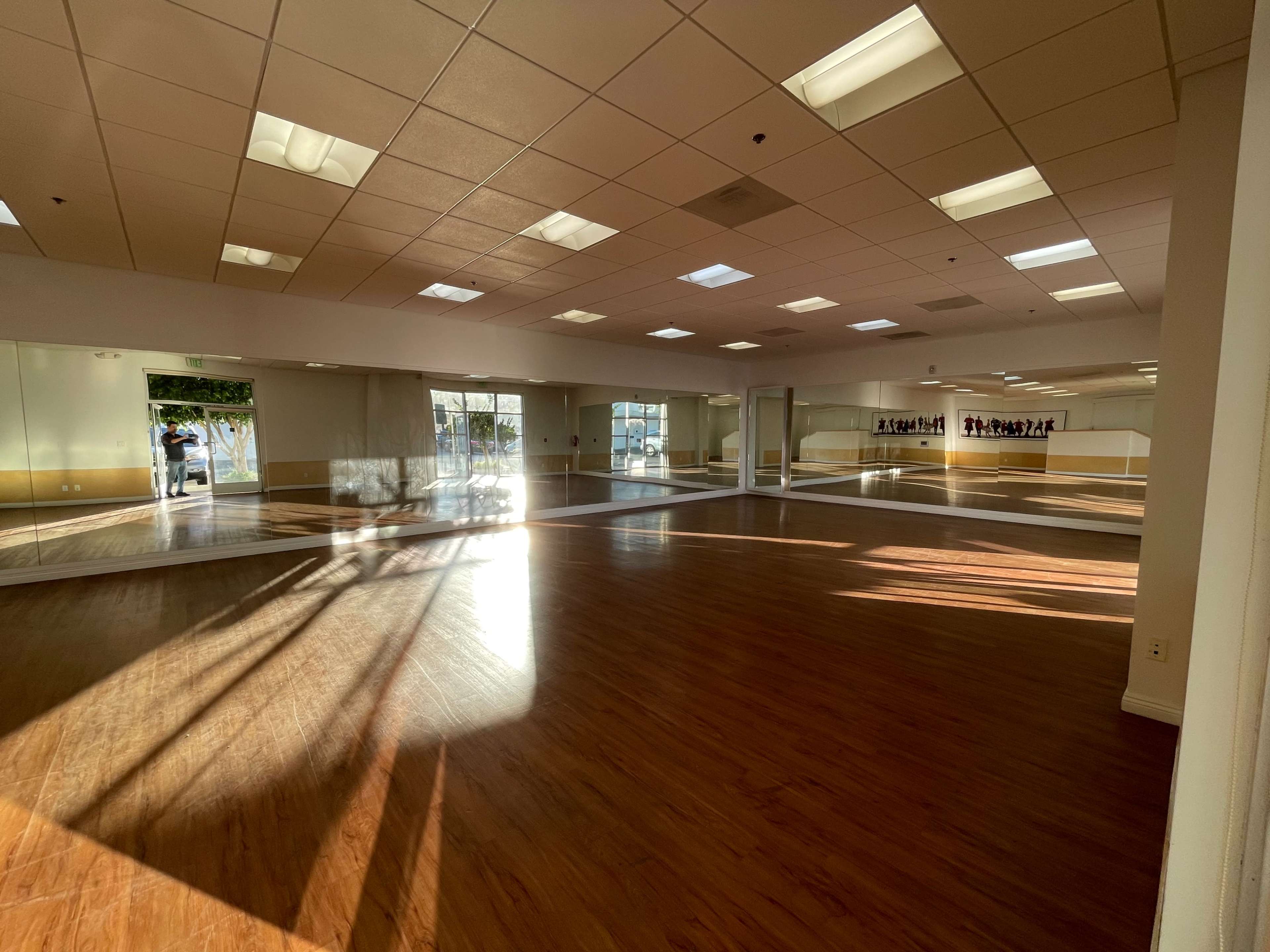 The image shows a spacious dance studio with wooden flooring and large mirrors on the walls, illuminated by natural light streaming through windows.