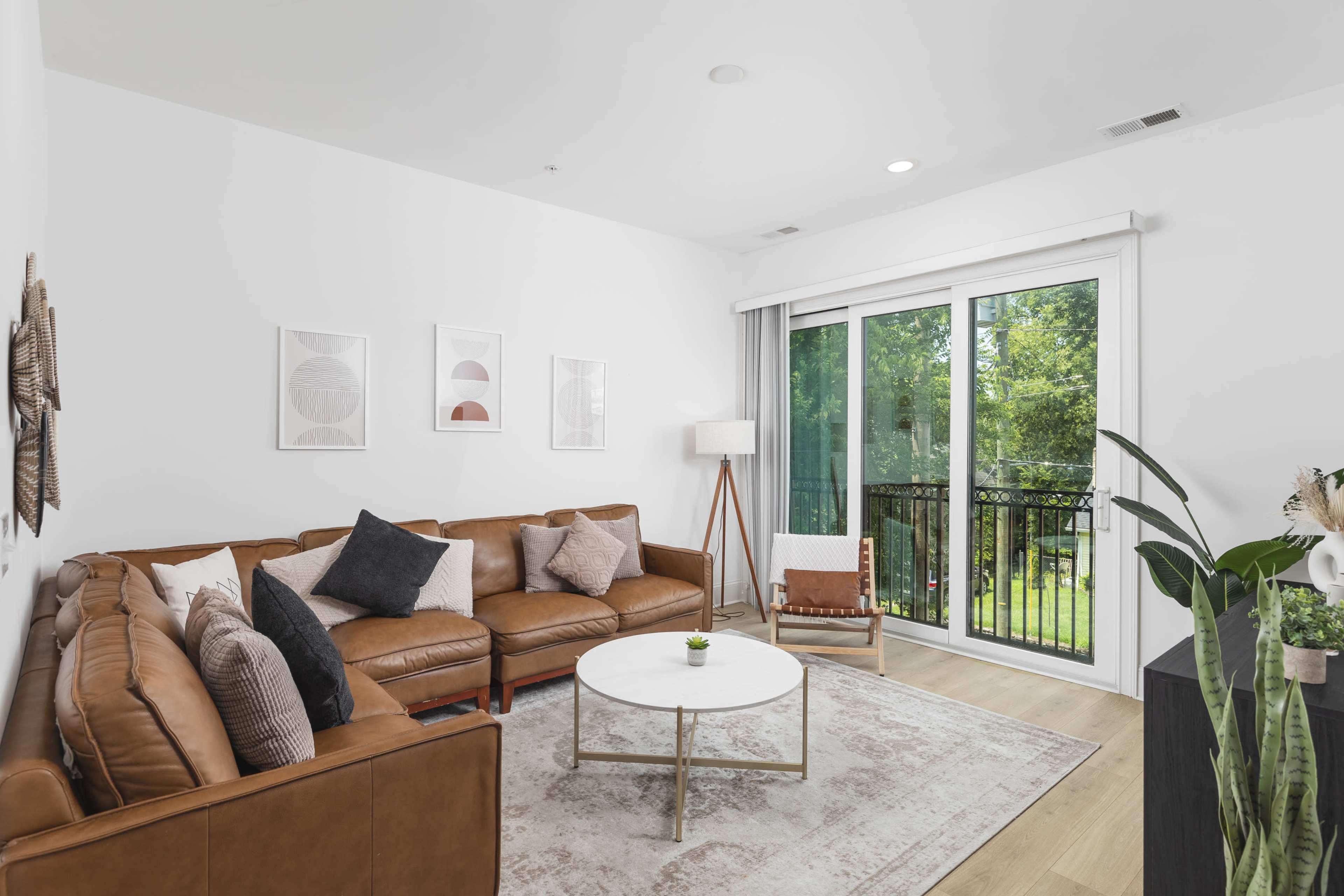 A modern living room features a brown leather sectional sofa, a round coffee table, and large windows that provide a view of greenery outside.