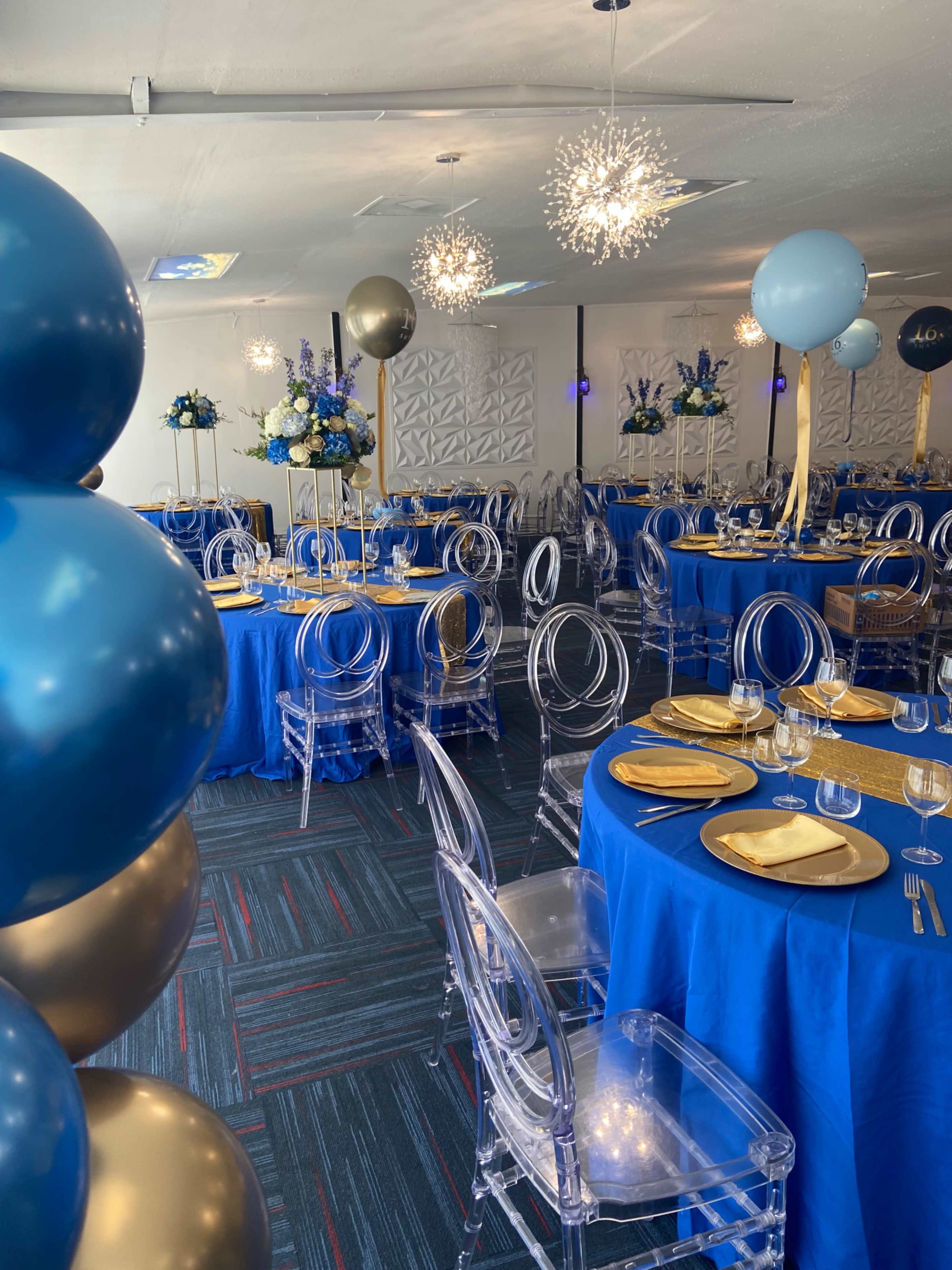 The image shows a decorated event space with blue tablecloths, gold accents, clear chairs, and floral arrangements, set for a gathering.