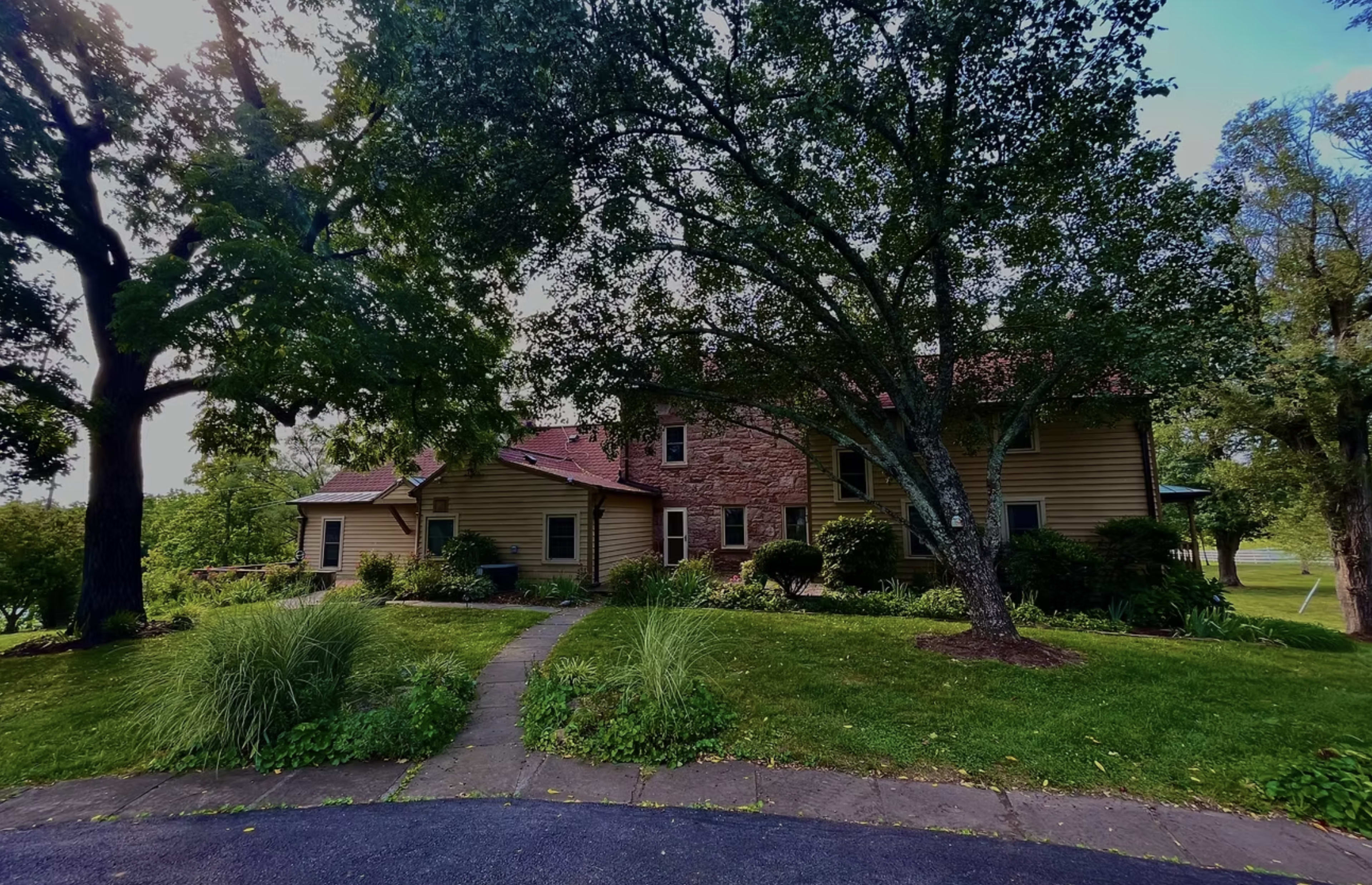 A large two-story house with a mix of brick and wooden siding is set amid lush greenery and shaded by mature trees.