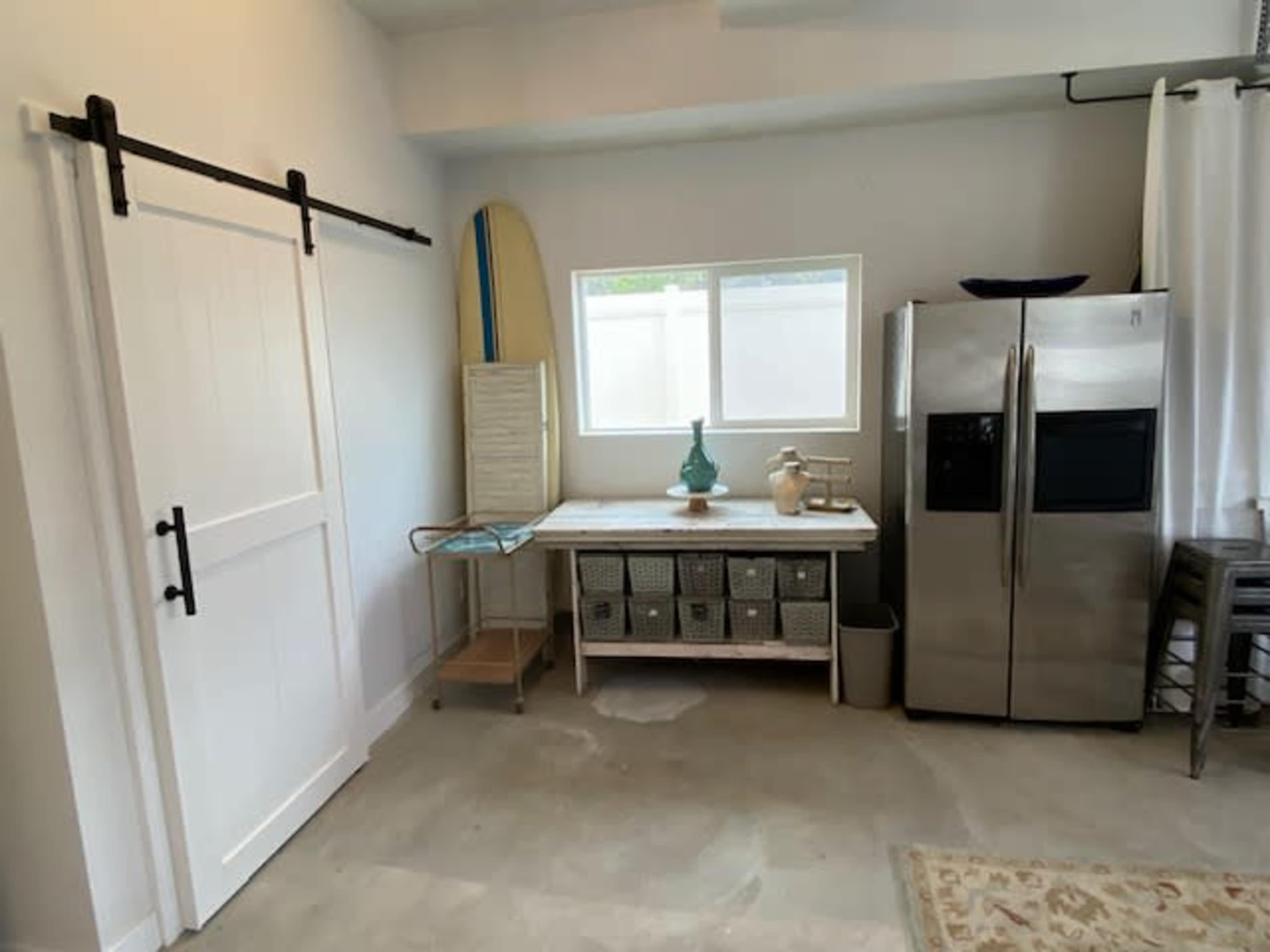 A room features a sliding barn door, a stainless steel refrigerator, a countertop with storage baskets, and a surfboard leaning against the wall.