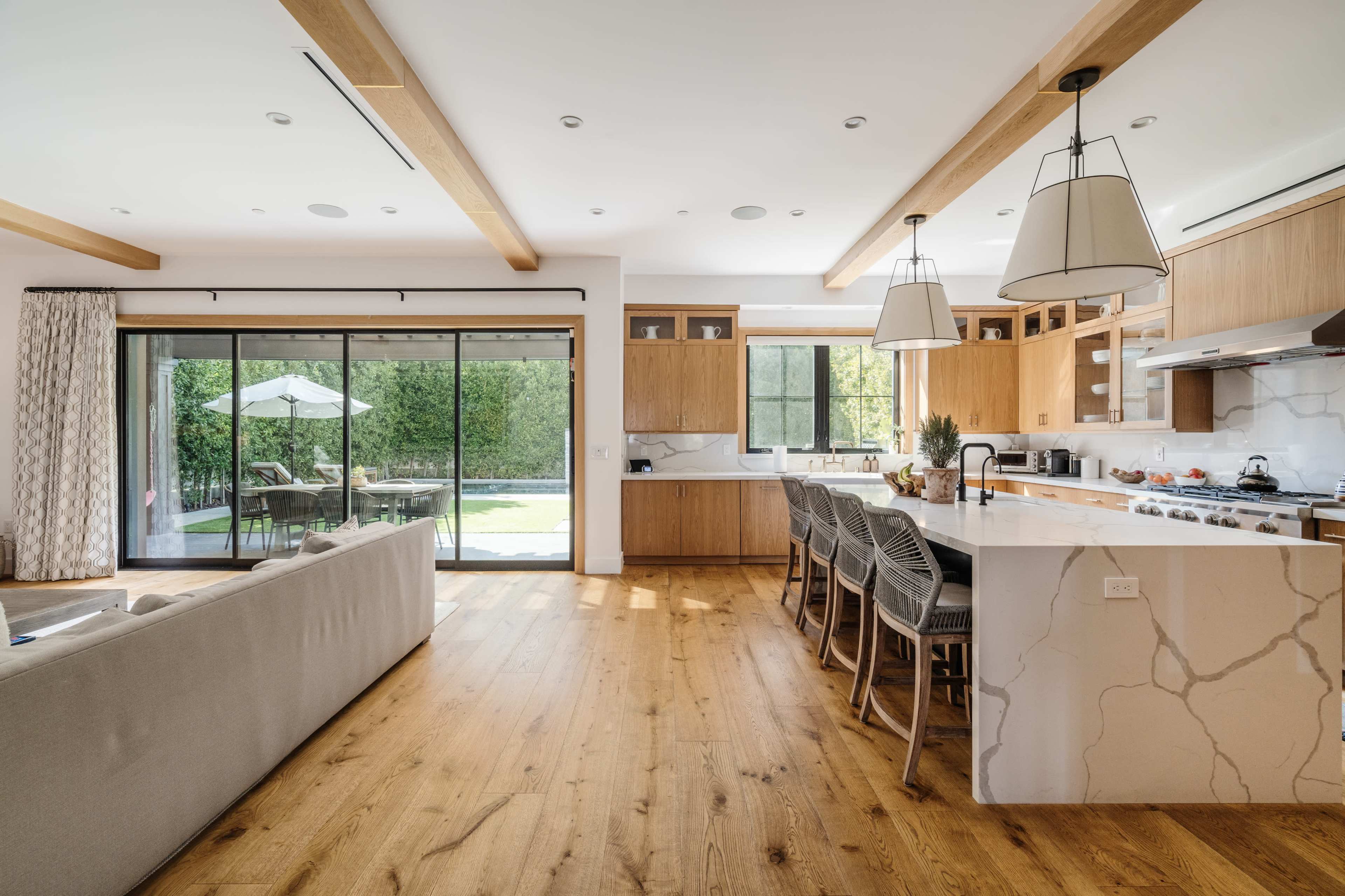 A modern kitchen and living area feature wooden cabinets, a large island with bar seating, and a view of an outdoor patio through glass sliding doors.
