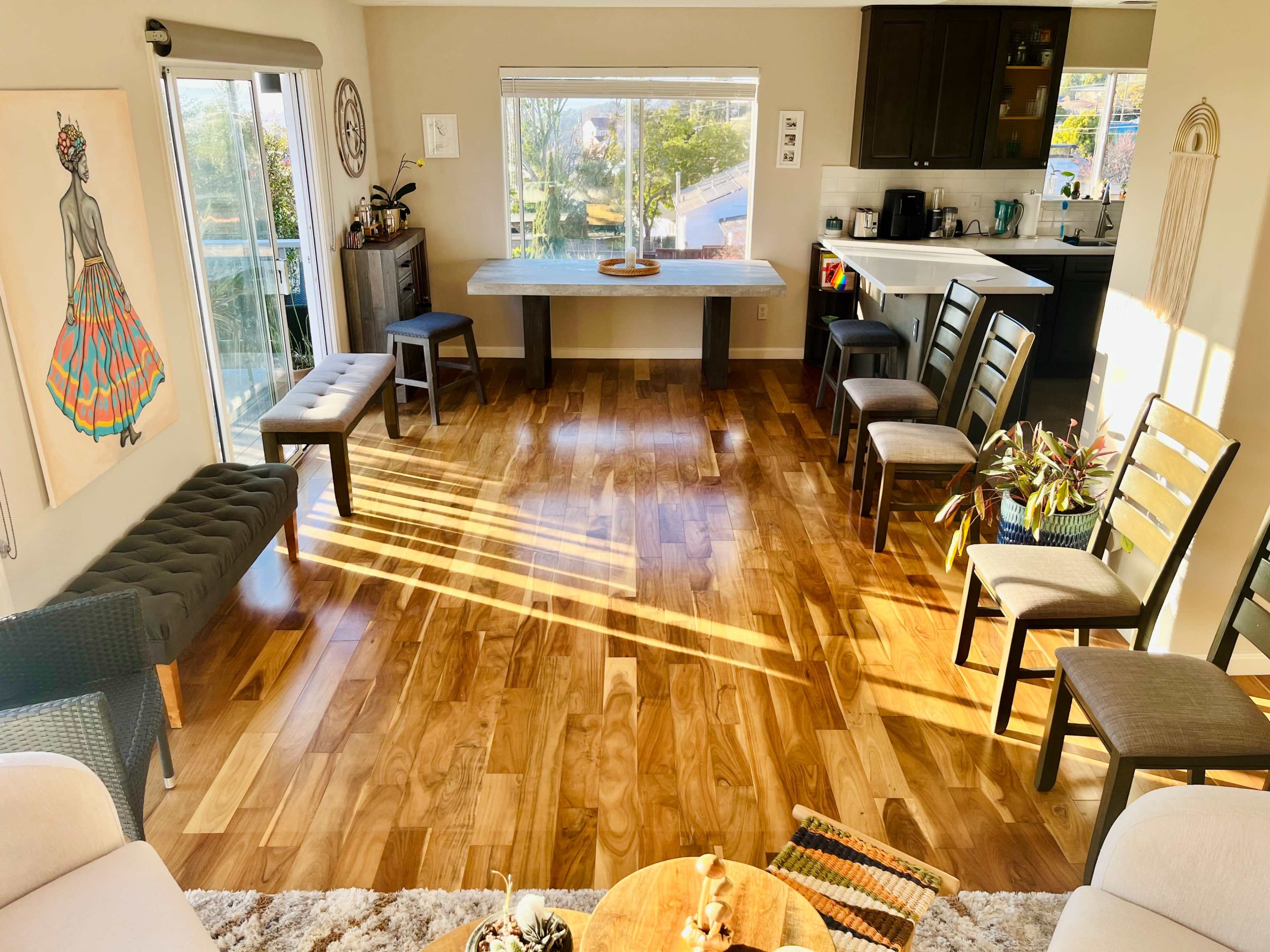 A spacious room features wooden flooring, a large table in the center, and chairs arranged around it, with large windows allowing natural light to illuminate the space.